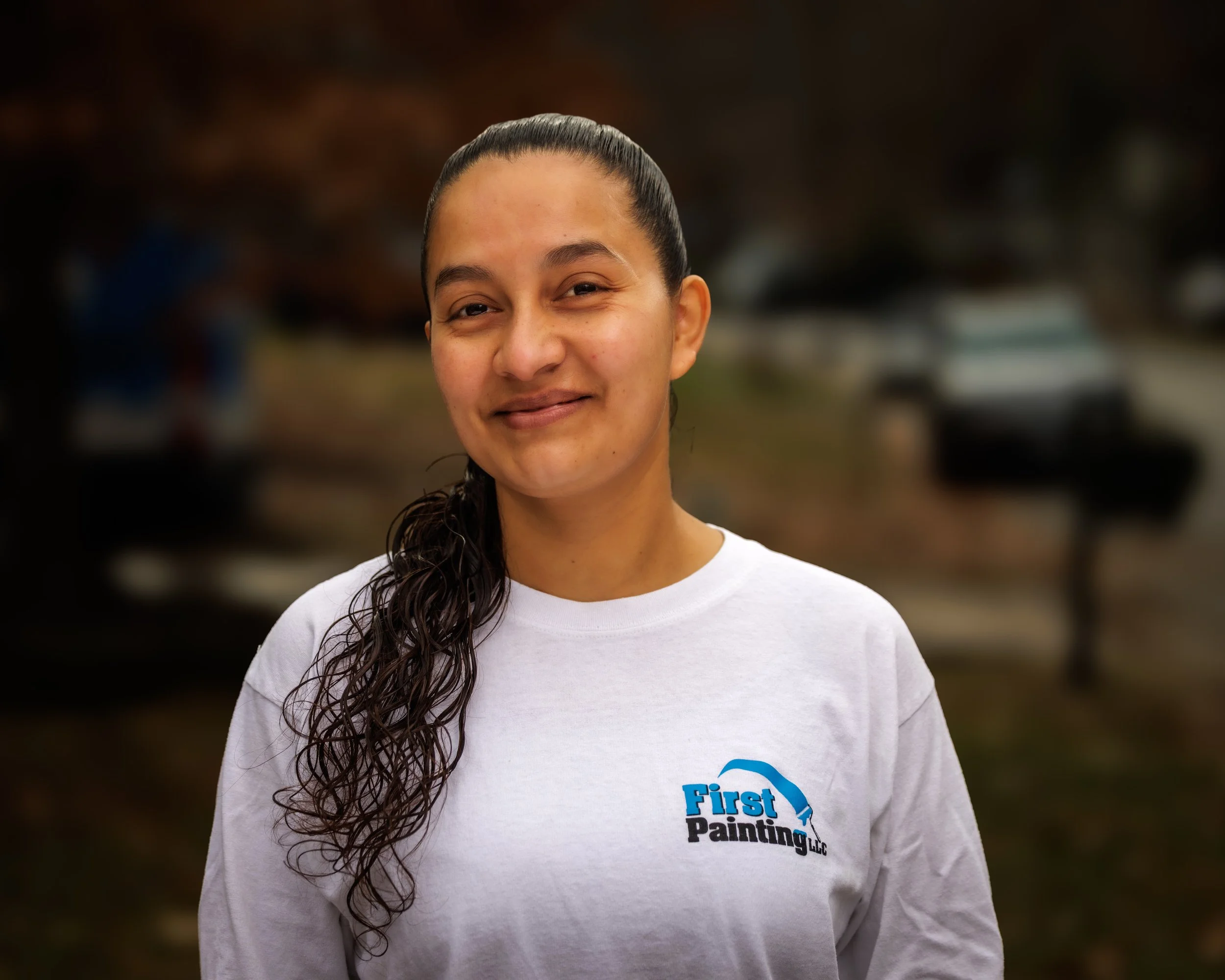 A woman with long, curly hair tied back, wearing a white T-shirt with a logo that says 'First Painting LLC,' standing outdoors with a blurred background of trees and a vehicle.