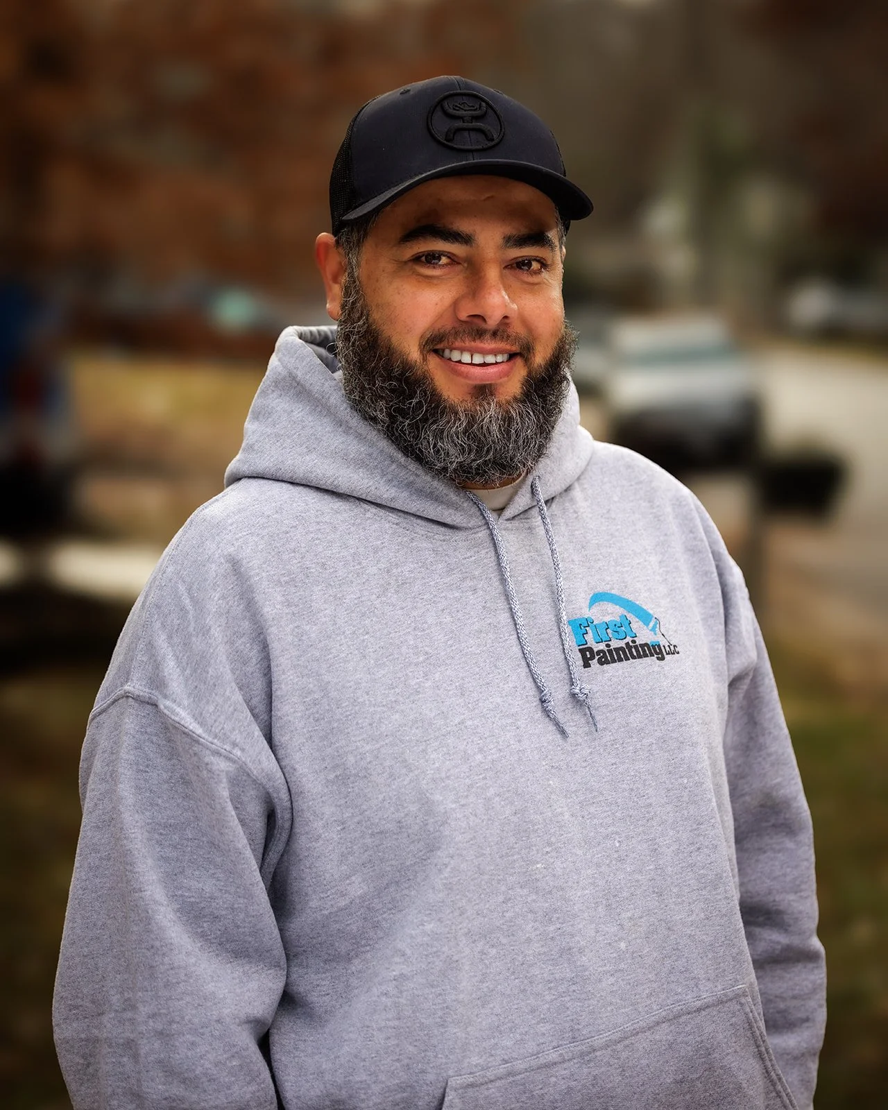A man with a beard smiling outdoors, wearing a gray hoodie with a logo and a black cap.
