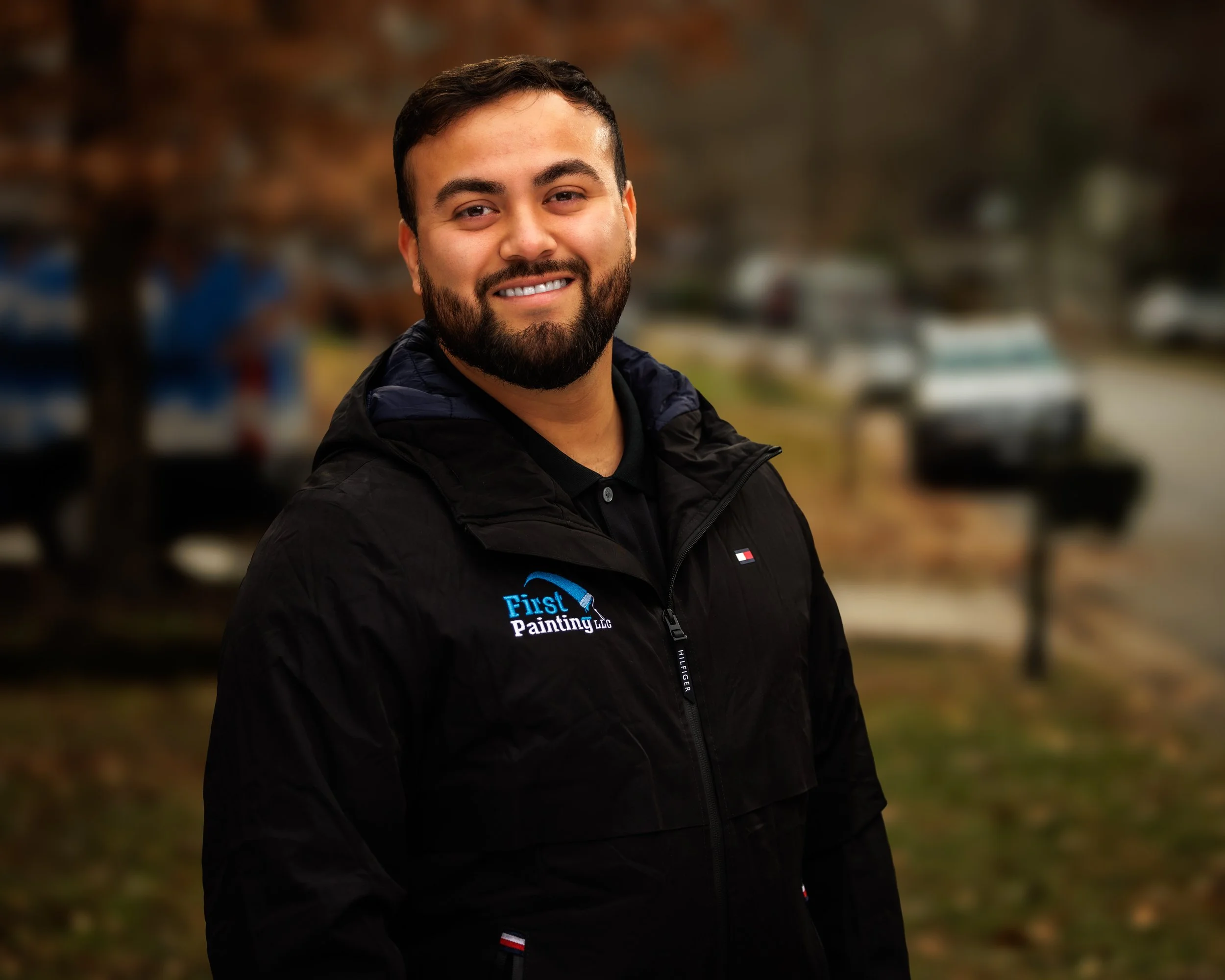 Smiling man standing outdoors wearing a black jacket with "First Painting LLC" logo, blurred trees and parked cars in the background.