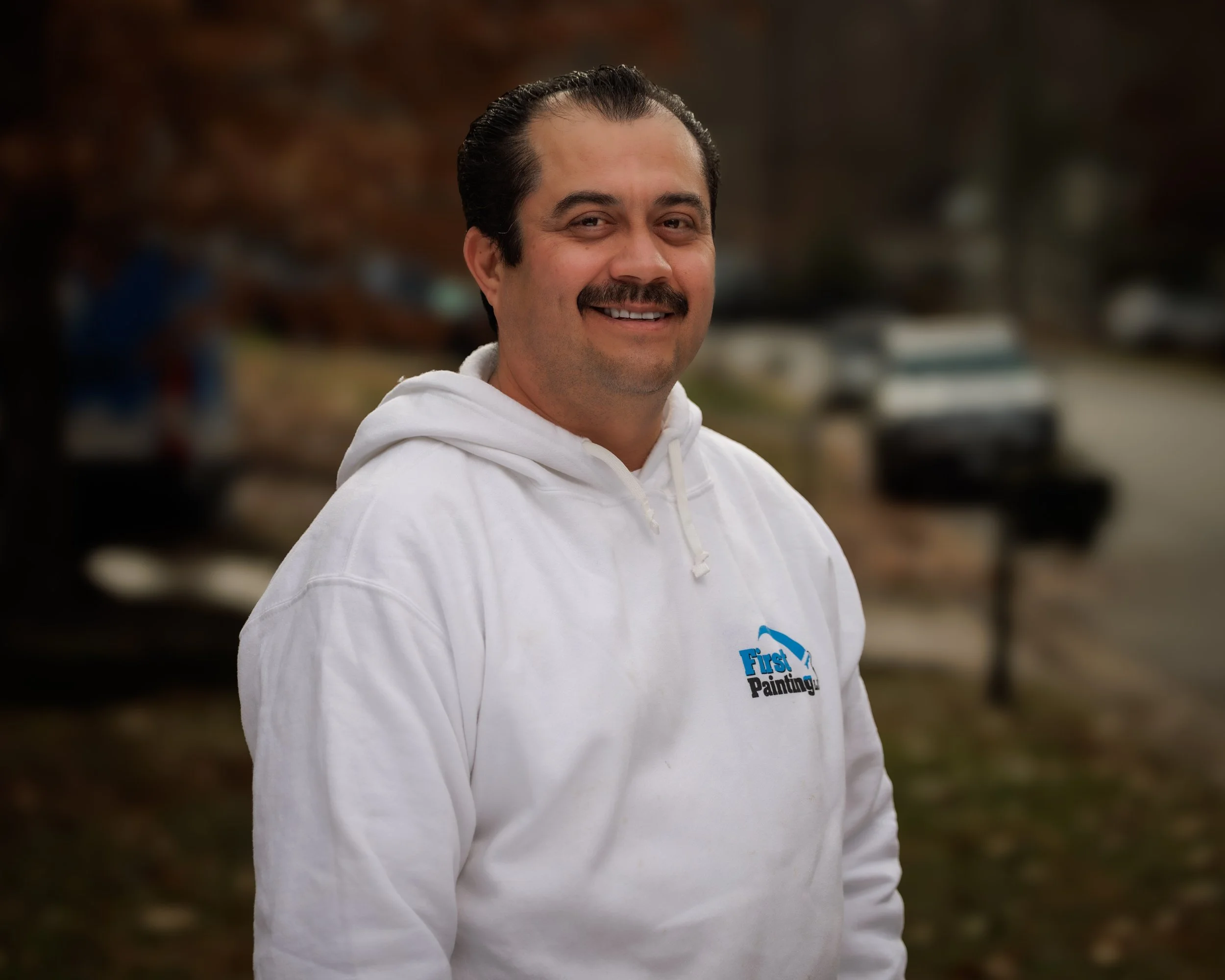 A man smiling outdoors wearing a white hoodie with a logo that says "First Painting". The background features parked cars and trees with fall foliage.