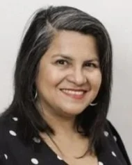 A woman with long dark hair smiling, wearing a black top with white polka dots, and hoop earrings.