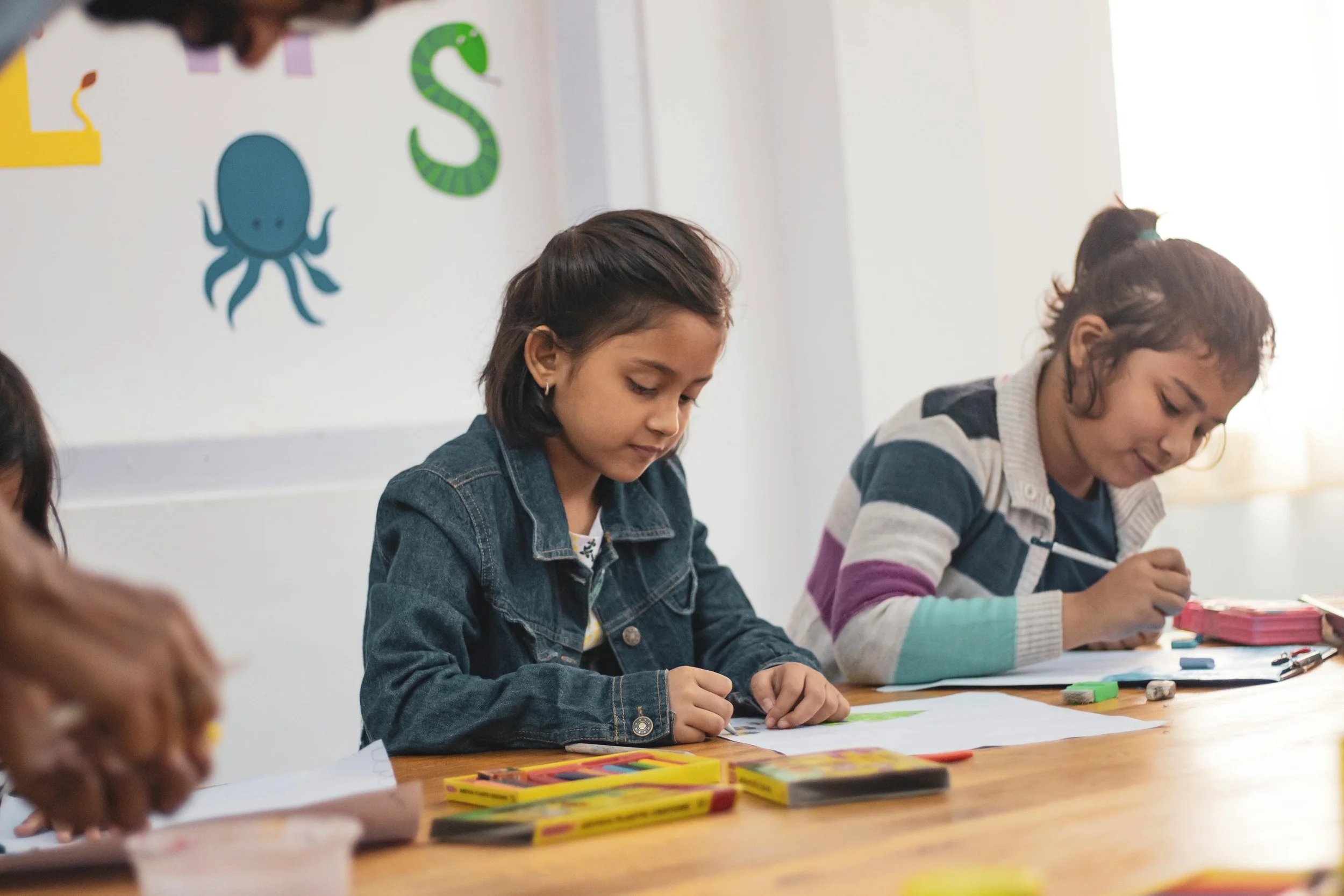 Two girls sitting at a wooden table, engaged in coloring or drawing with markers and papers, in a classroom with colorful wall decorations including an octopus and alphabet letters.