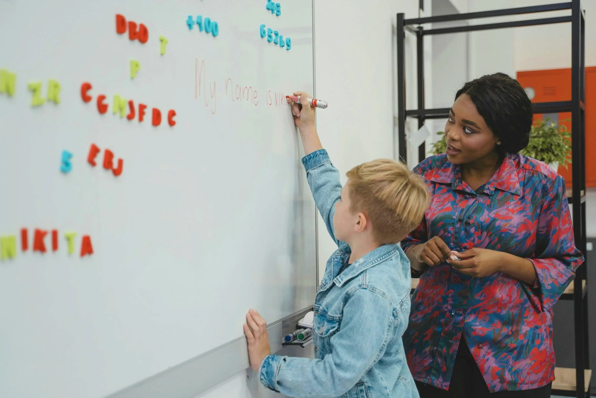 A teacher assists a young student writing on a whiteboard in a classroom. The whiteboard has colorful magnetic letters and numbers, and the boy is writing a sentence. The teacher looks on with a supportive expression.