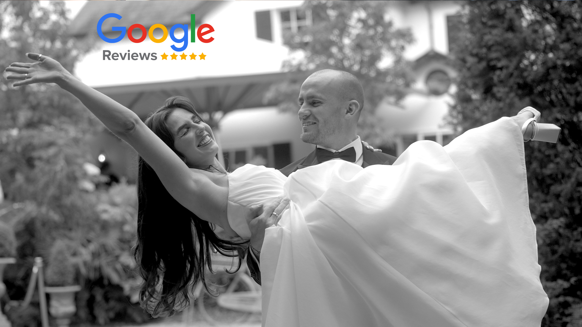 A groom in a tuxedo holding a smiling bride in a wedding dress, dancing outdoors in front of a house with trees and a patio umbrella in the background.