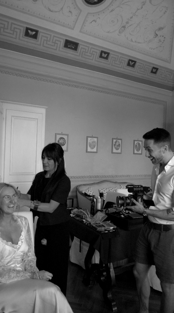 The bride at bridal prep with Eye Flower Films' videographer Breffny. One woman is sitting, another woman is standing applying makeup or adjusting something, and Breffny is holding the camera with a smile as a wedding moment is captured.