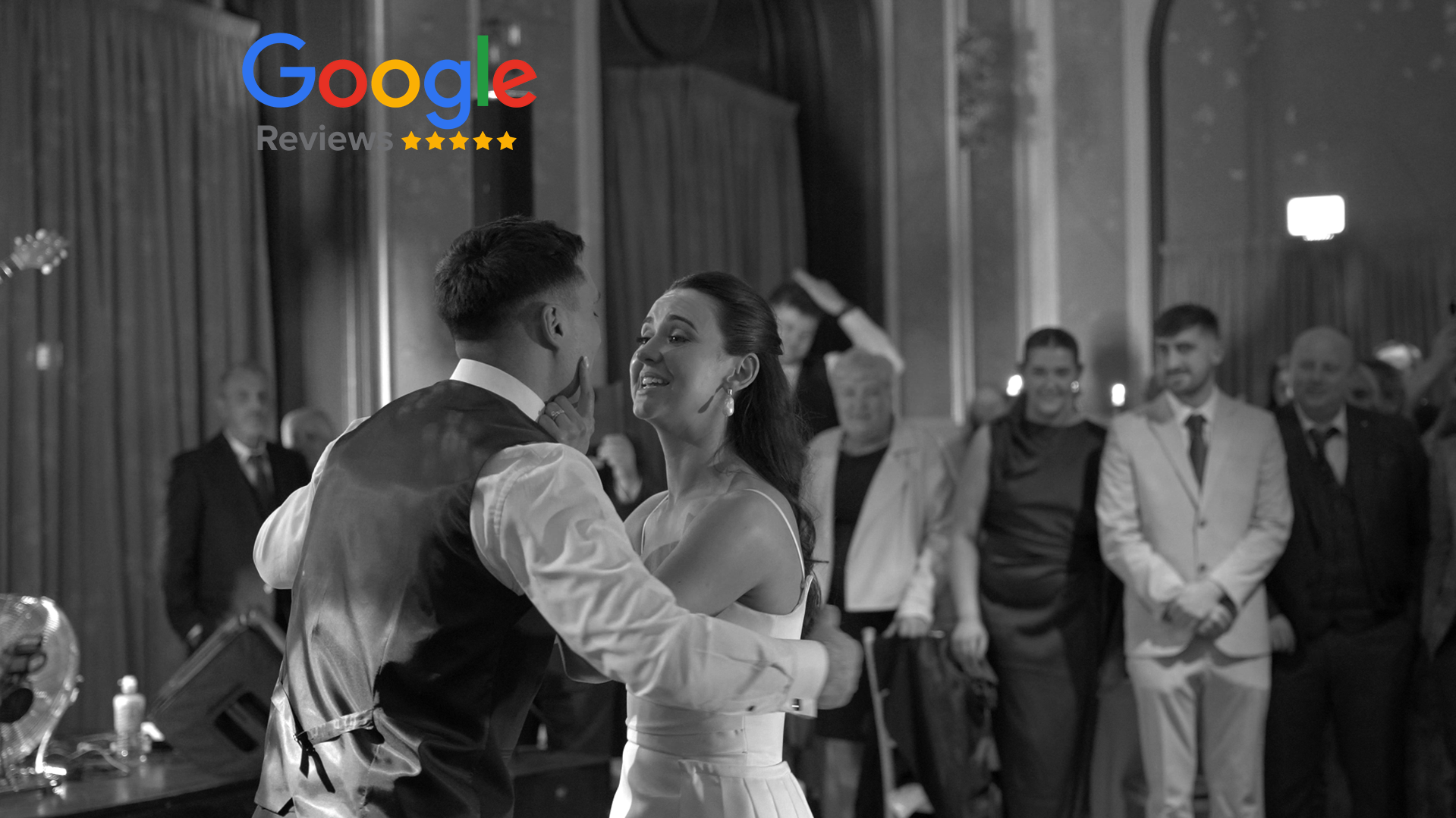 A black and white photo of a couple dancing closely at a formal event, surrounded by onlookers dressed in suits and dresses.