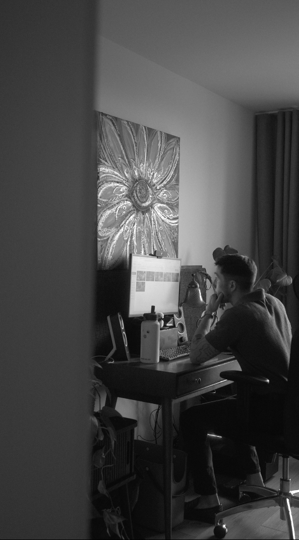 Breffny, the owner of Eye Flower Films at his editing desk in his home office, using a computer with a large monitor, surrounded by speakers and personal items, with Eye Flower Films' painting on the wall.