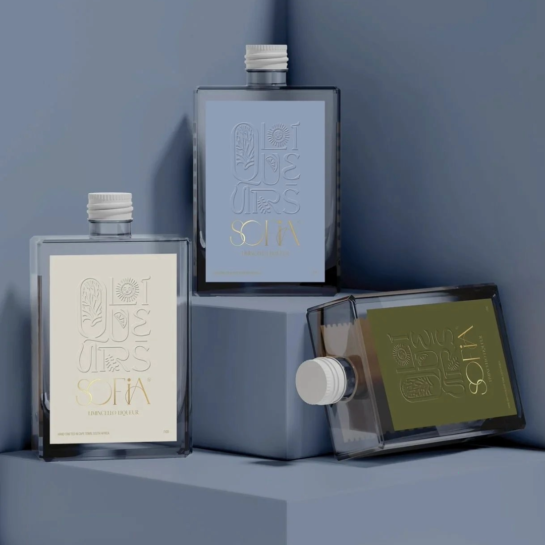 Three bottles of SOFIA limited edition liquor with minimalist labels, two upright and one lying on its side, on a modern blue surface and wall background.