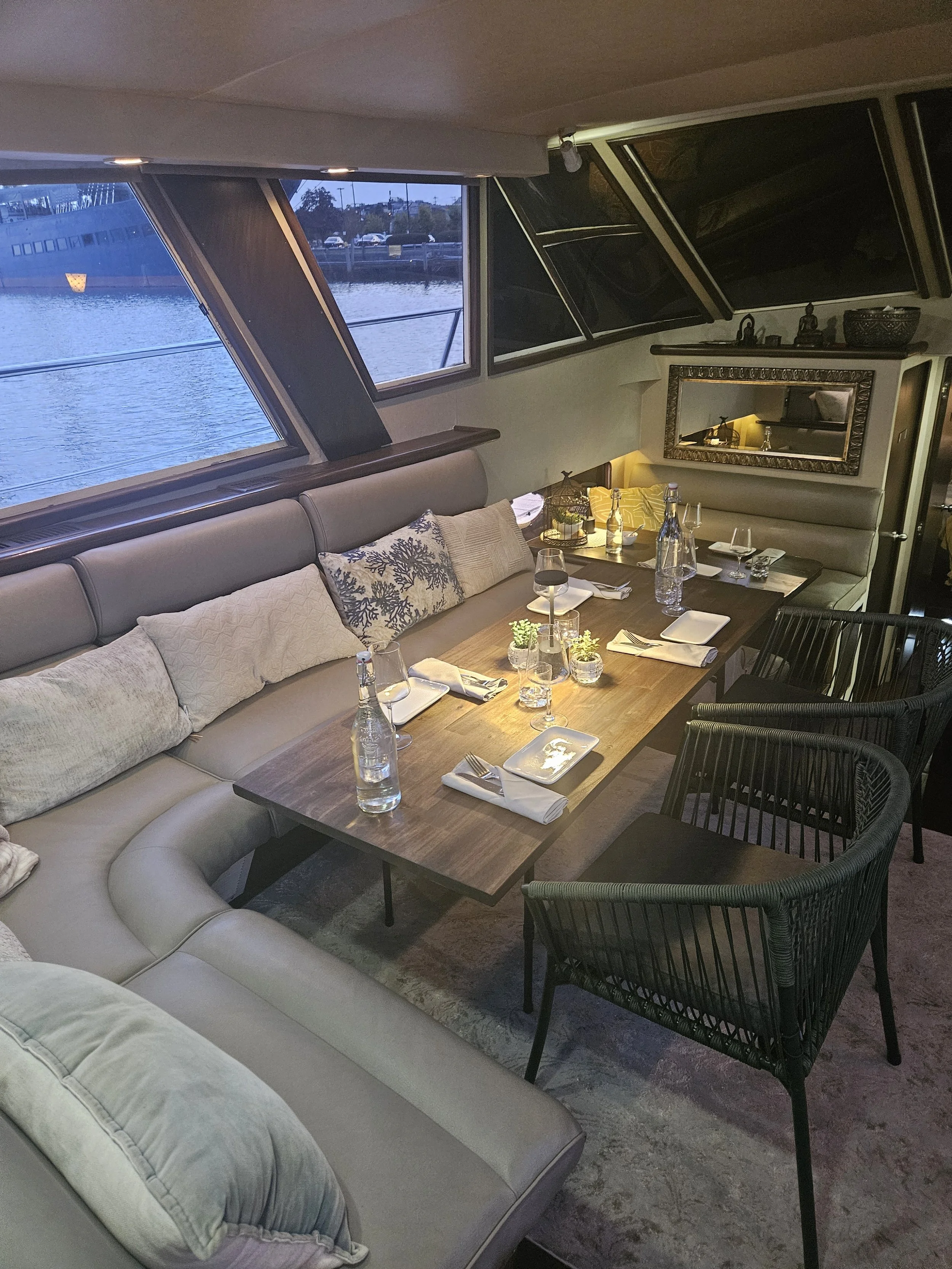 Interior of a boat with large windows showing water and a shoreline outside. The room features a long beige curved sofa with decorative pillows, a wooden dining table set with wine glasses, water bottles, napkins, and small plants, and black woven chairs surrounding the table. A mirror and decorative items are on the far wall.