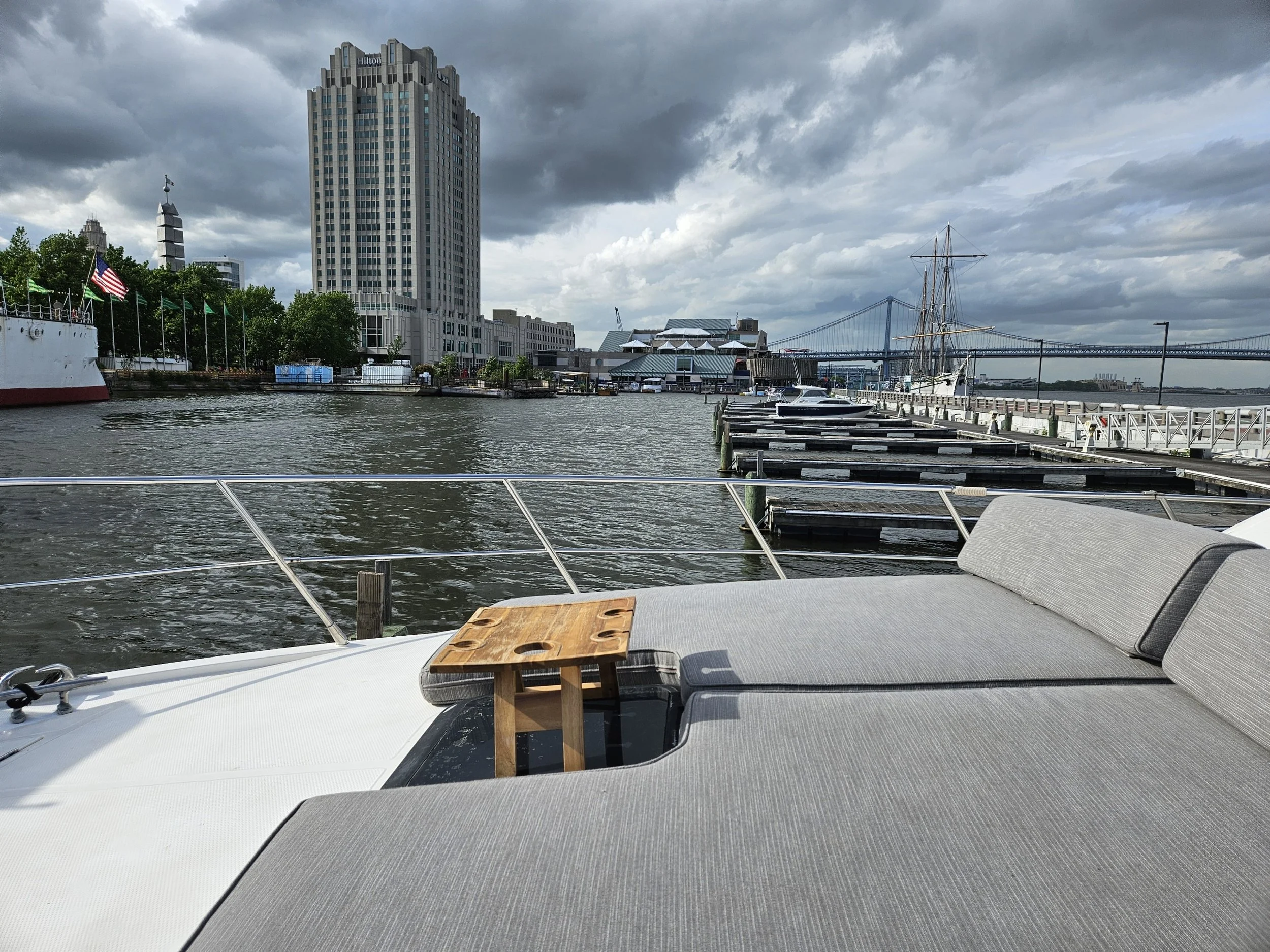 View from a boat showing a dock with boats, a tall Hilton hotel building, and a bridge in the background under cloudy sky.