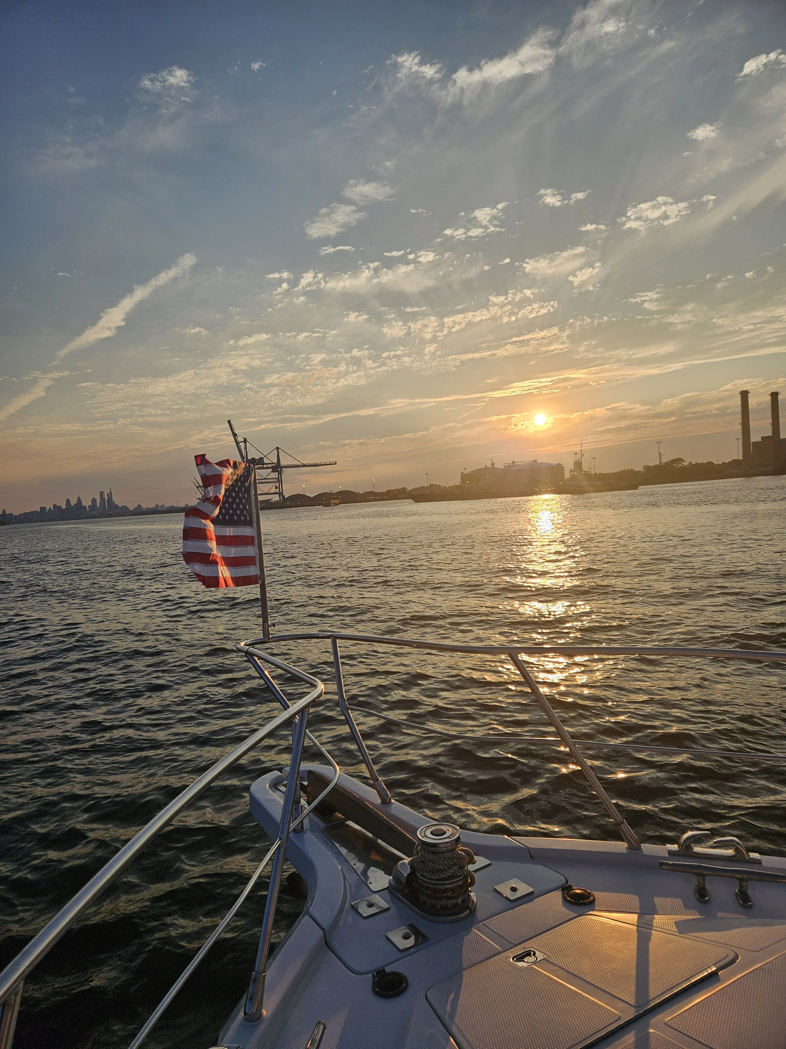 View from a boat showing a sunset over a city skyline, with some clouds in the sky and an American flag on the boat's railing.