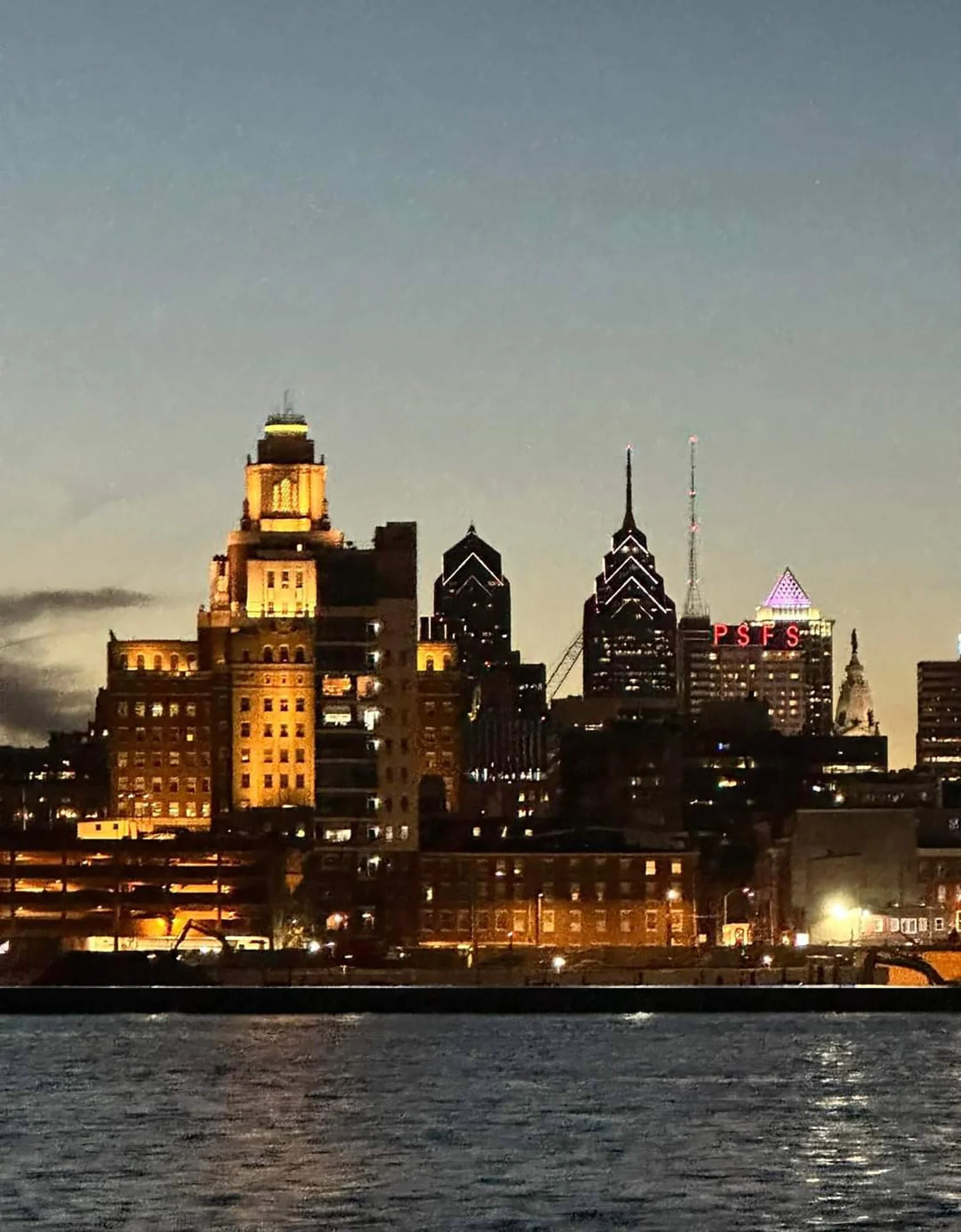 Night city skyline with illuminated tall buildings, some with lit-up signs, viewed over a dark body of water.
