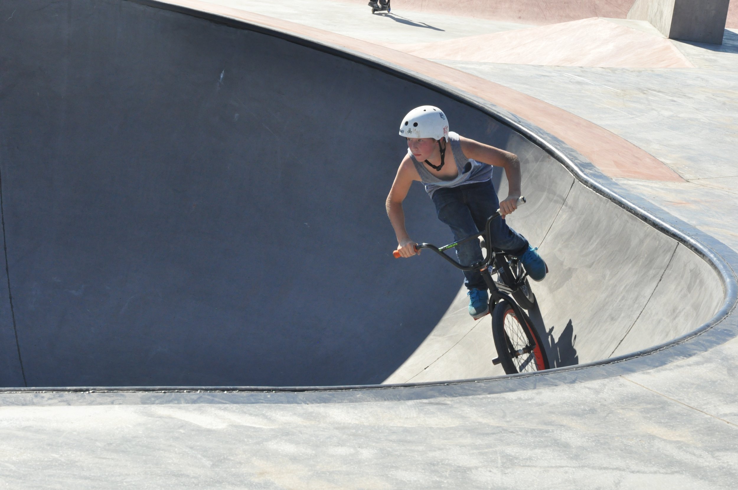 A young boy wearing a white helmet rides a BMX bike in a skatepark bowl with gray concrete walls and a curved edge.