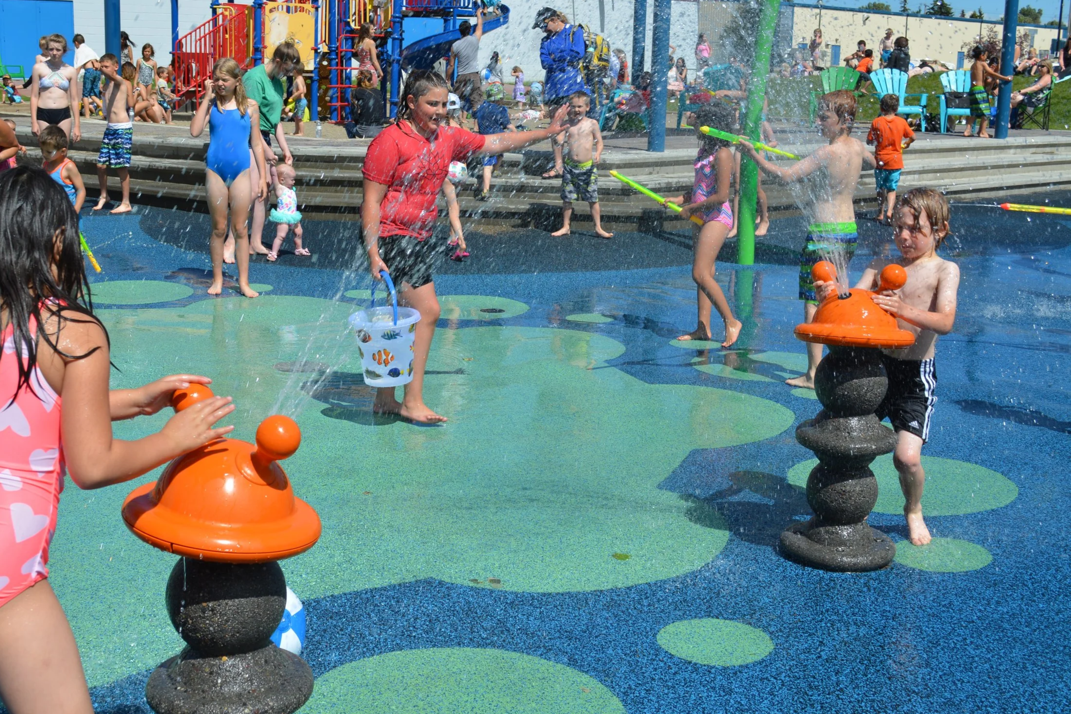 Children playing and splashing in a water park on a sunny day, with many kids and adults in the background.