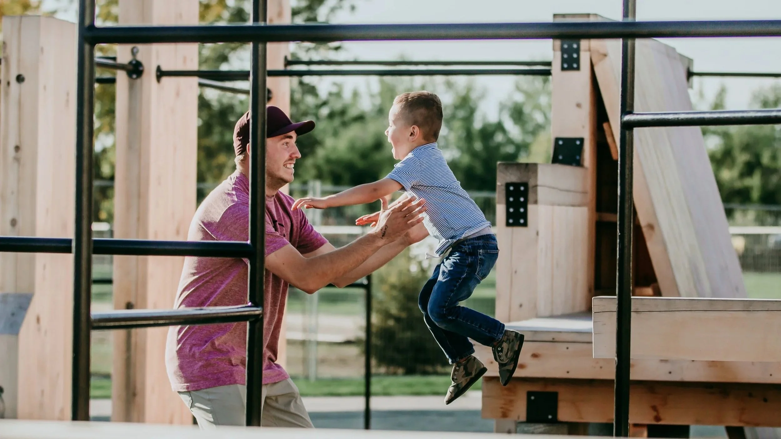 A man and a young boy playing on a wooden playground structure outdoors, with the man holding the boy mid-air as they smile at each other.