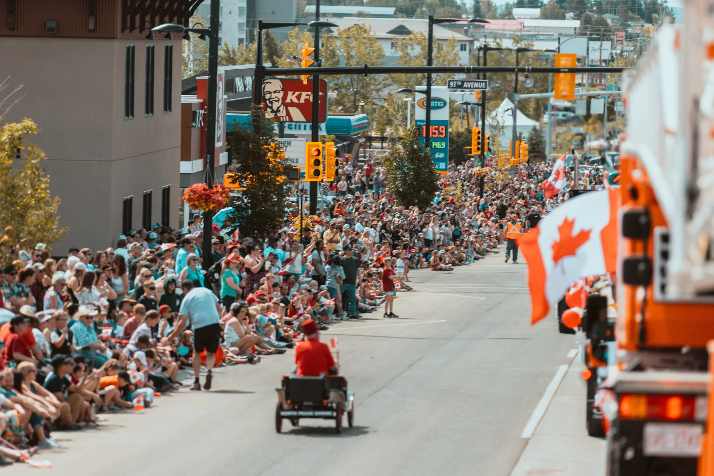 Crowd of people sitting and standing along a street during a parade, with some holding Canadian flags, and parade vehicles, including one with a Canadian flag, moving down the street.