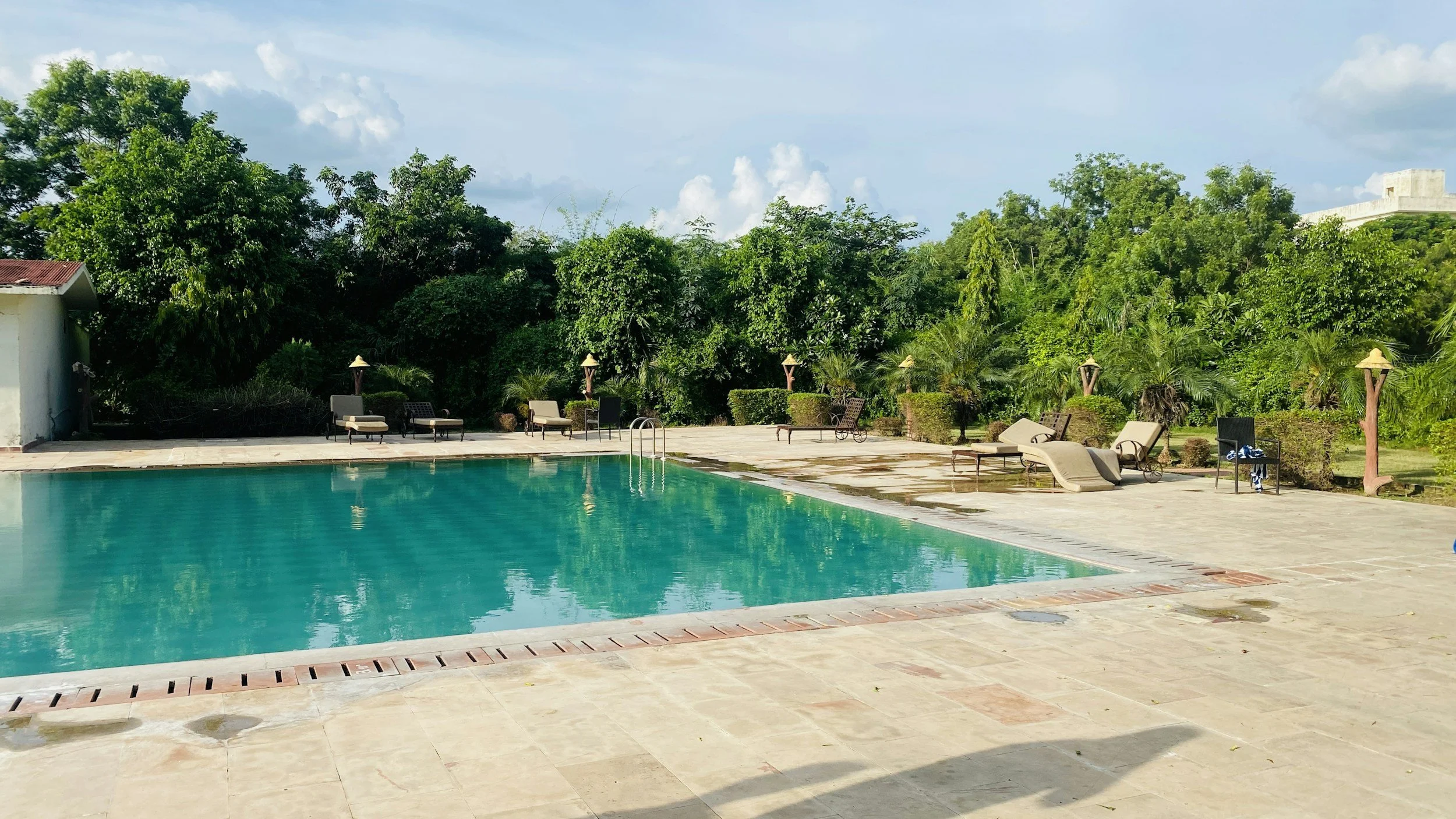 An outdoor swimming pool surrounded by lounge chairs, umbrellas, and lush green trees under a partly cloudy sky.