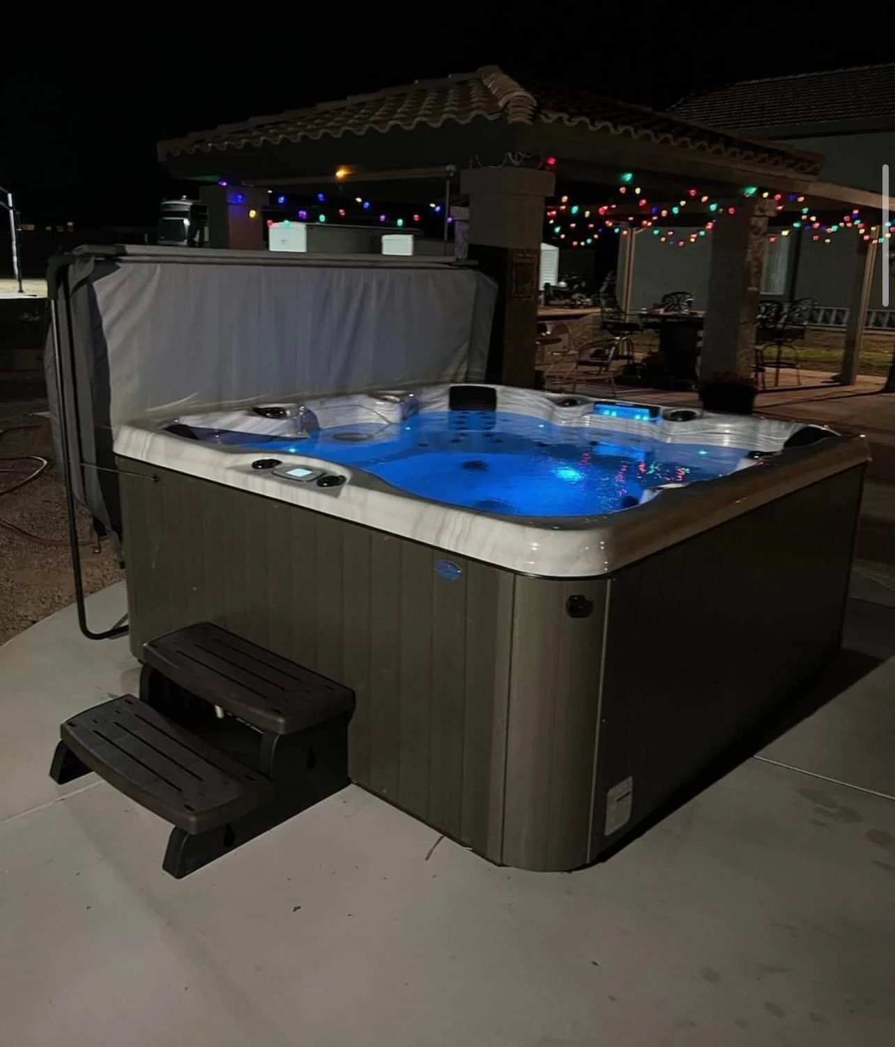 Nighttime outdoor shot of a hot tub with blue-lit water, illuminated controls, and a set of small stairs, with a covered patio decorated with string lights in the background.