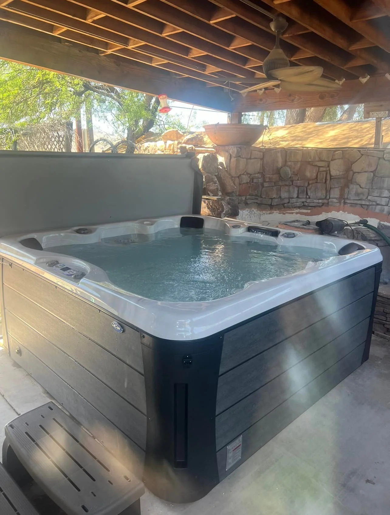 An outdoor hot tub on a patio, with a stone wall and trees in the background, under a wooden roof.