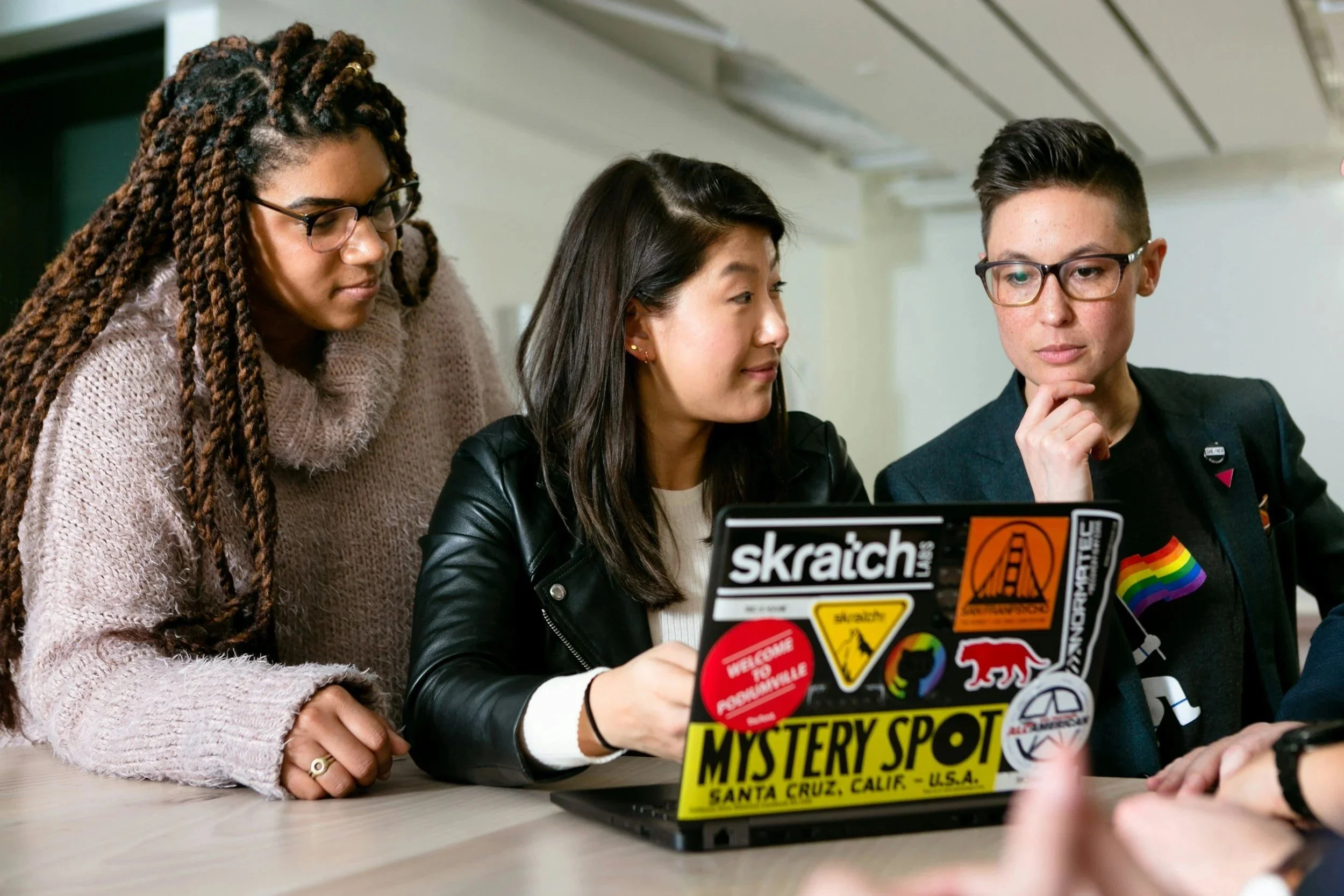 Drei vielfältig aussehende Personen schauen auf einen Laptop, der auf einem Tisch steht, mit Stickerdekoration. Sie scheinen gemeinsam an einem Projekt zu arbeiten.
