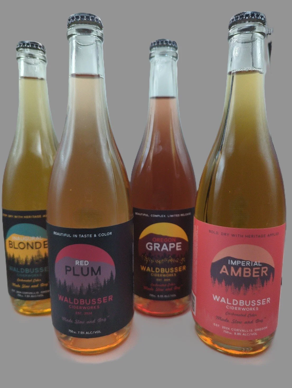 Four glass bottles of cider with colorful labels, including flavors like Red Plum, Grape, Amber, and Blonde, from Waldbusser Ciderworks.