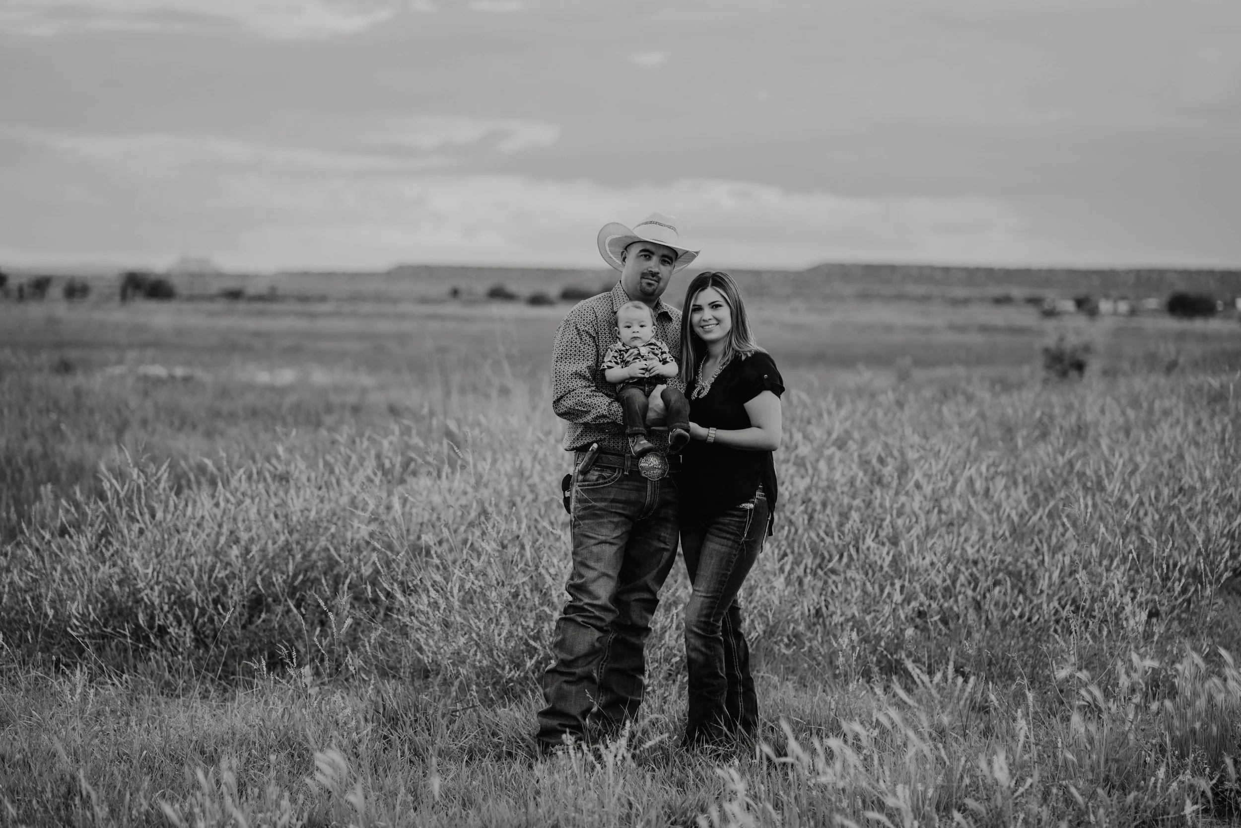 A man, woman, and baby standing together in a grassy field under a cloudy sky, with the man wearing a cowboy hat and the woman and baby smiling.