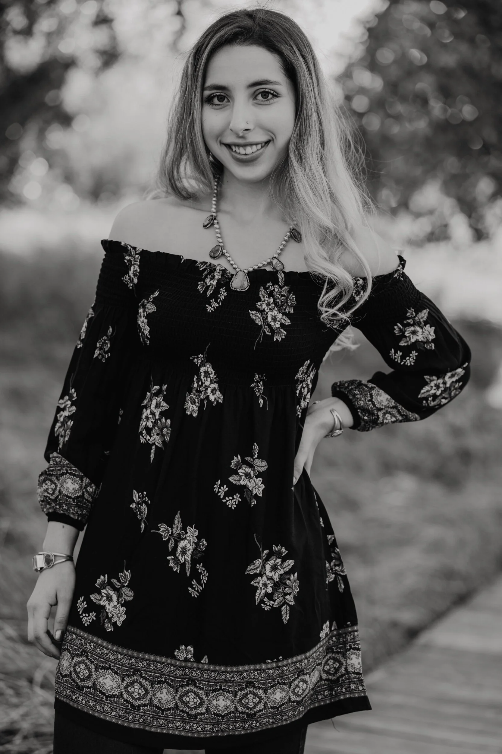 Black and white photo of a young woman with long, wavy hair, smiling, wearing an off-the-shoulder floral dress and a necklace, standing outdoors.