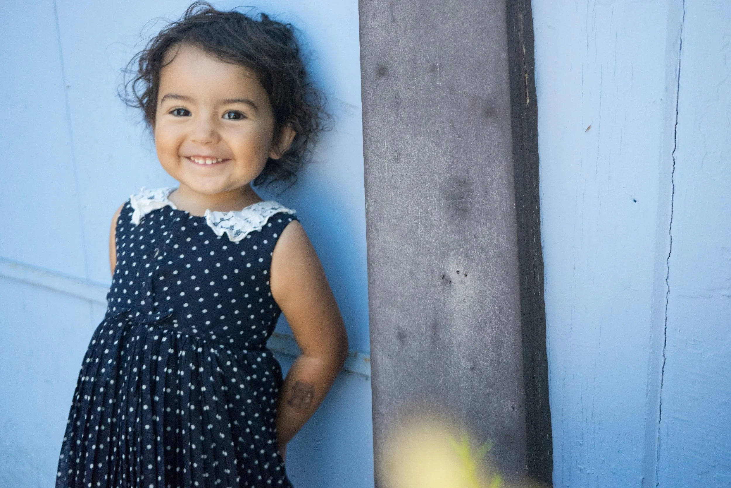 A young girl with curly dark hair, smiling and standing against a light blue wall and a concrete column, wearing a navy blue polka dot dress with a lace collar.