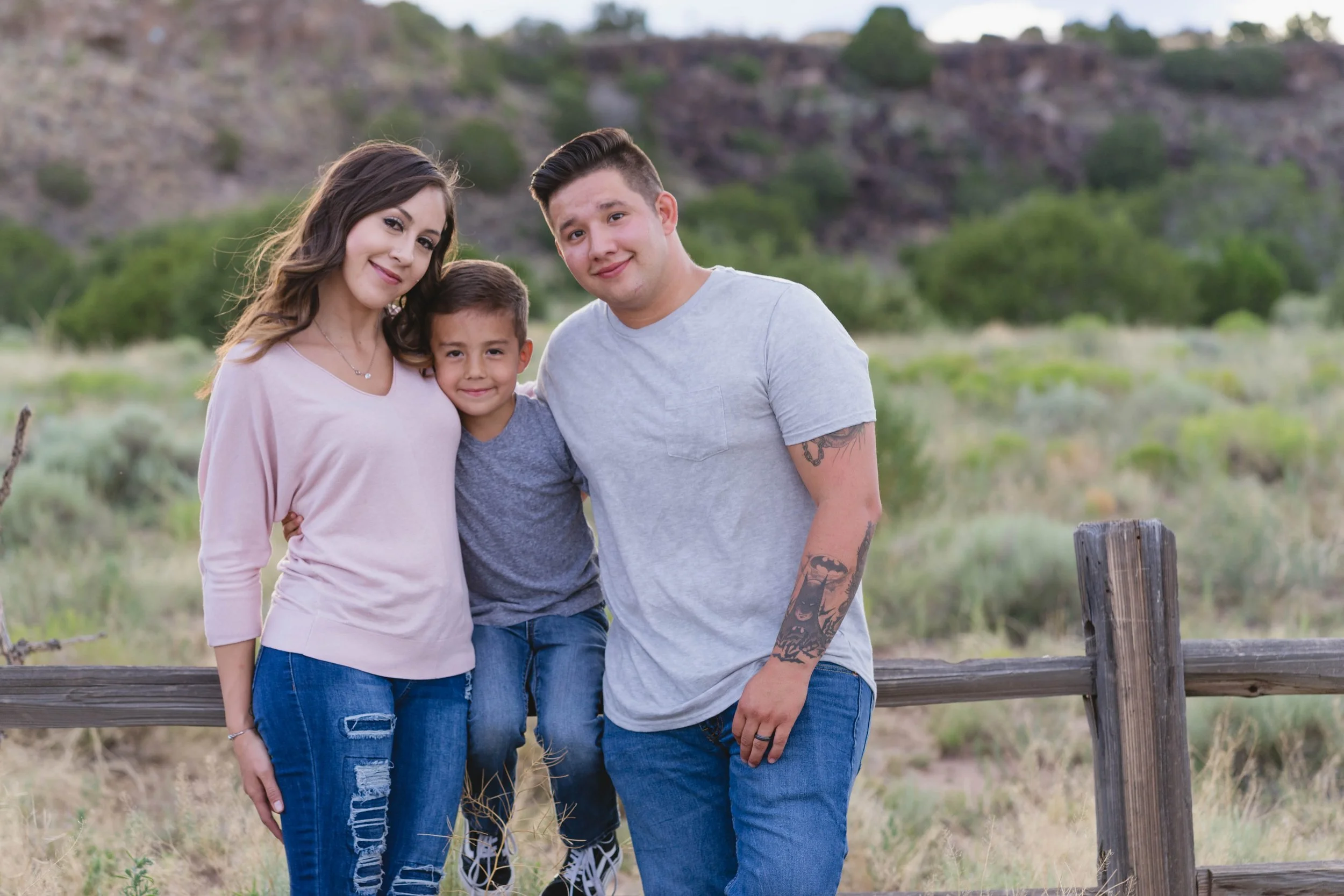 A family of three standing outdoors near a wooden fence, smiling at the camera, with green trees and hills in the background.