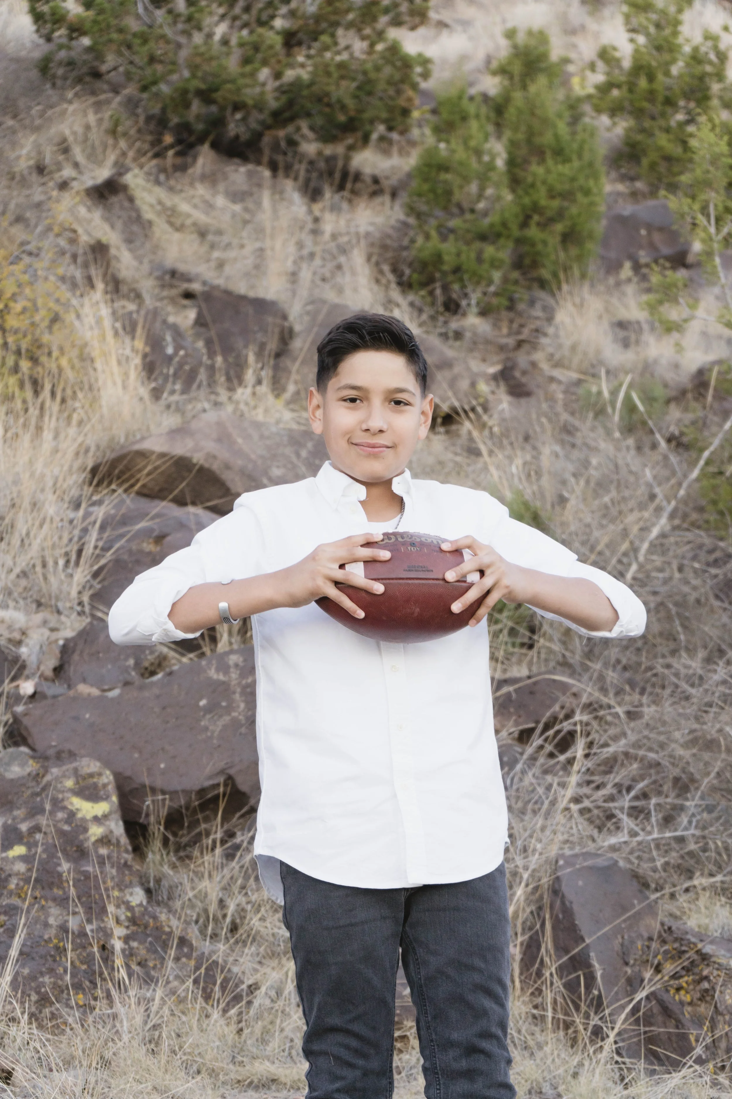 A young boy with dark hair, wearing a white button-up shirt and dark jeans, standing outdoors on rocky terrain with dry grasses and trees in the background, holding a football with both hands.