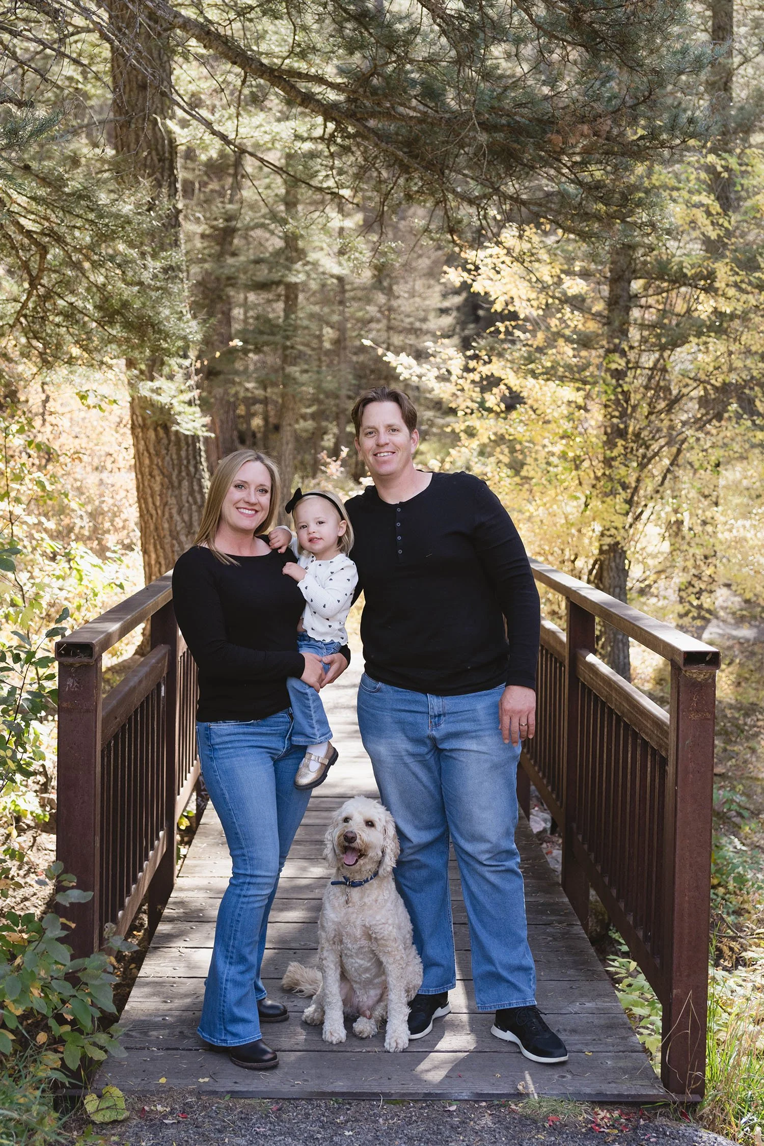 A family of three and a dog standing on a wooden bridge in a forest, smiling at the camera in autumn.