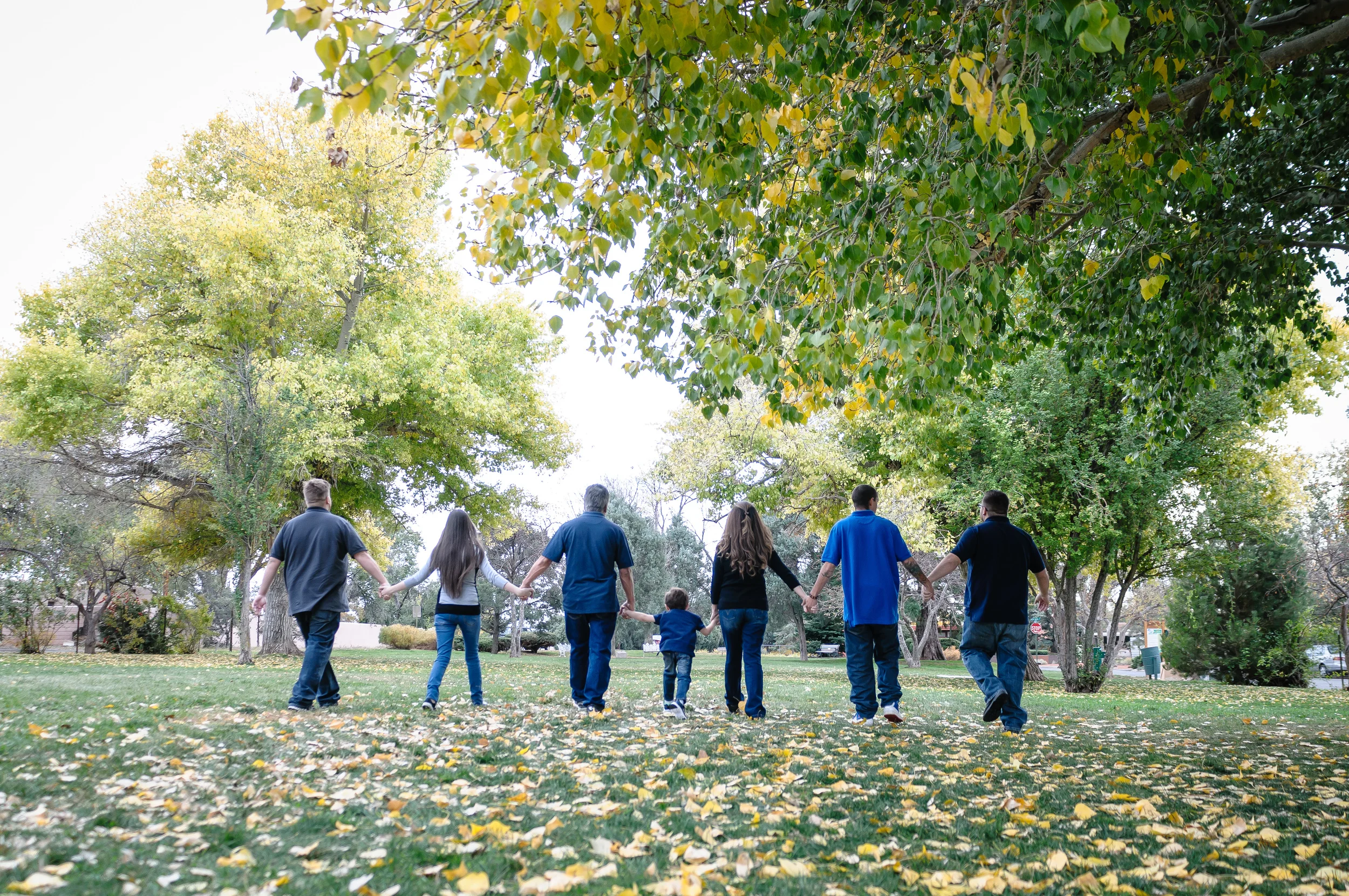 A group of seven people, including children and adults, holding hands and walking together across a park with fallen leaves and green trees.