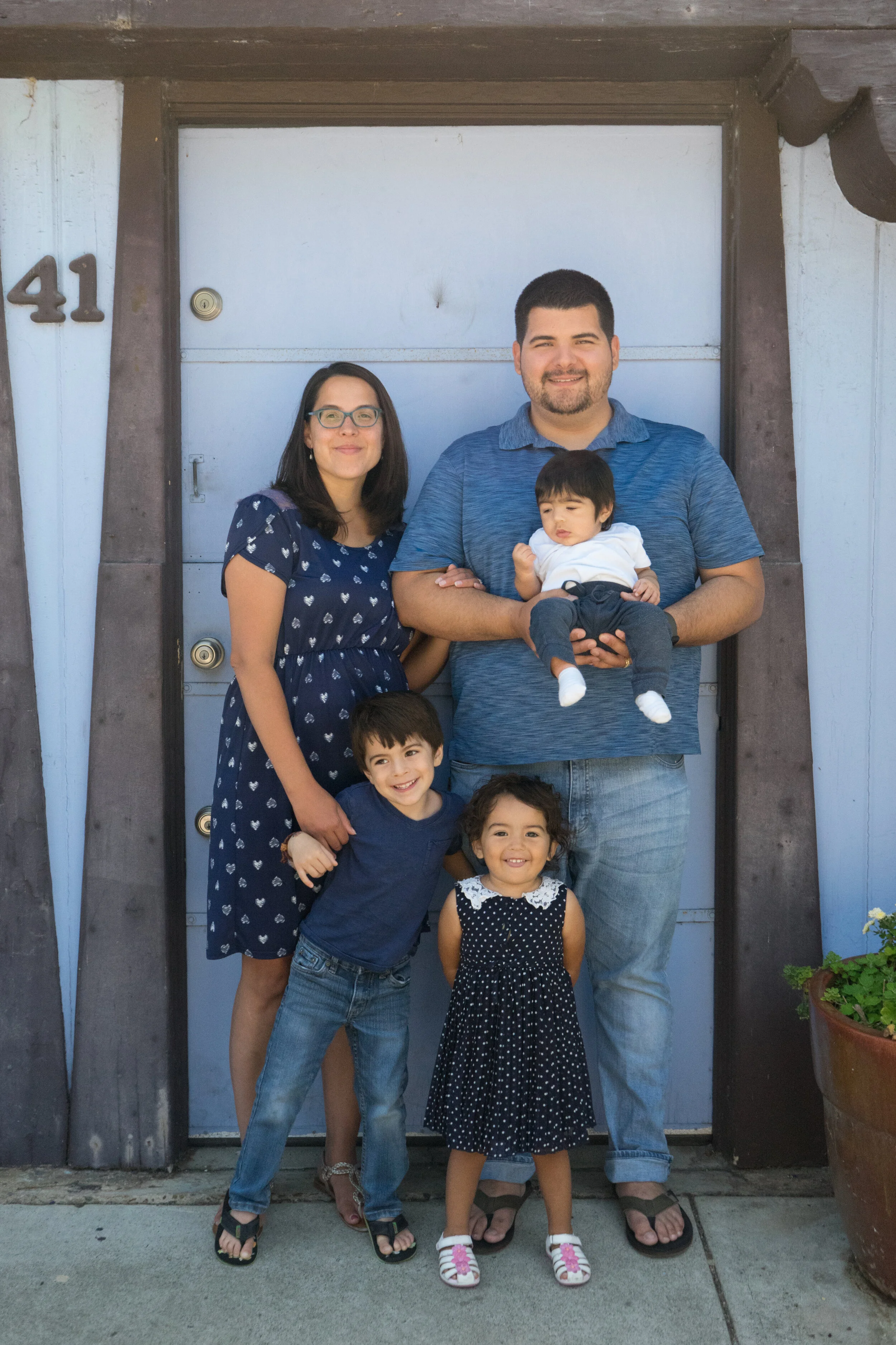 A family of six standing in front of a house door, including a woman wearing glasses, a man holding a baby, two young children, a boy, and a girl, all smiling.