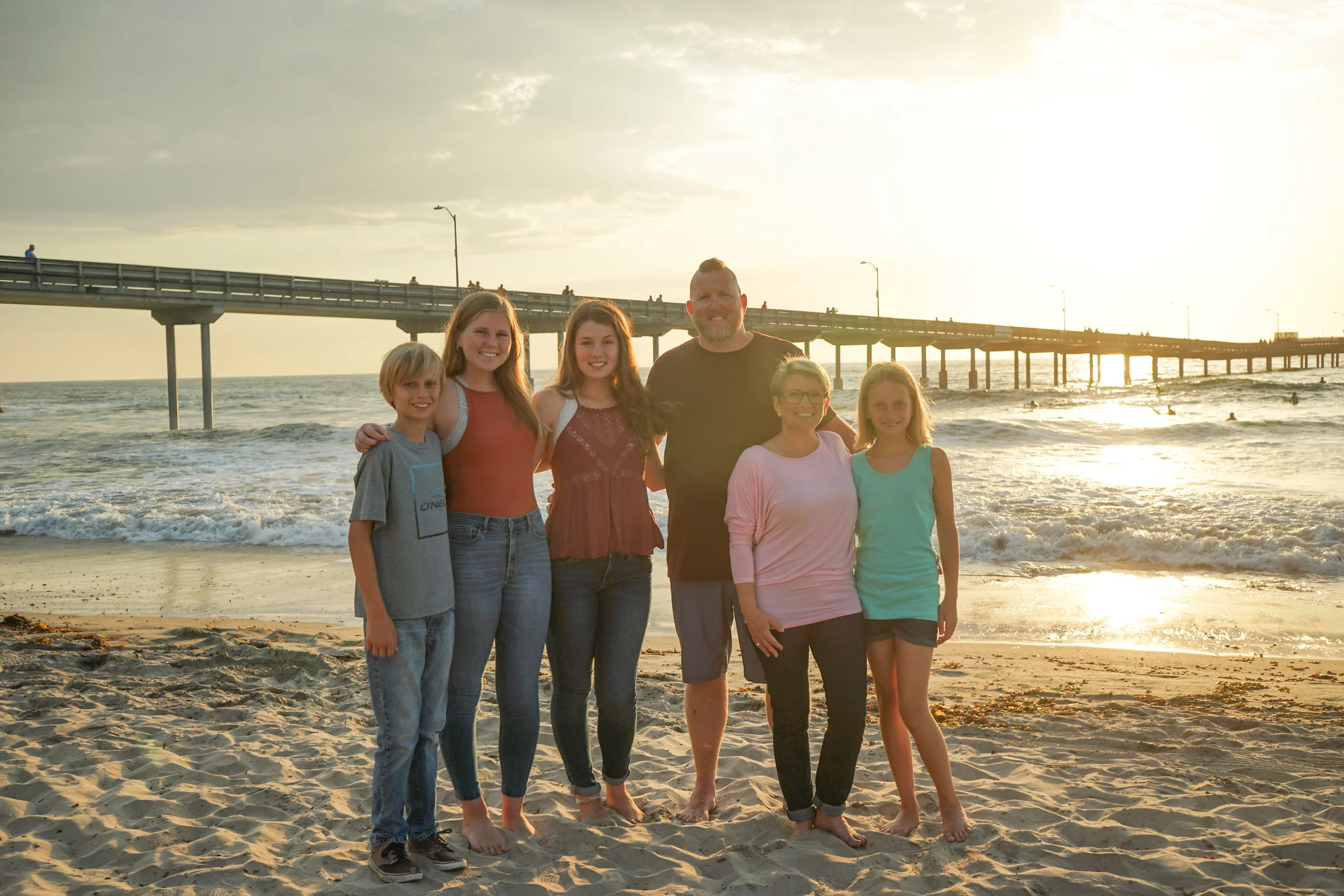 A family of six smiling and posing on the beach at sunset, with a pier extending into the ocean in the background.