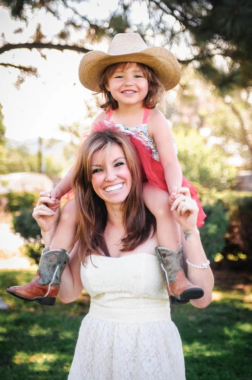 A woman with long brown hair smiling, carrying a young girl on her shoulders, who is wearing a large sun hat and a red dress, in a park with trees and sunlight.