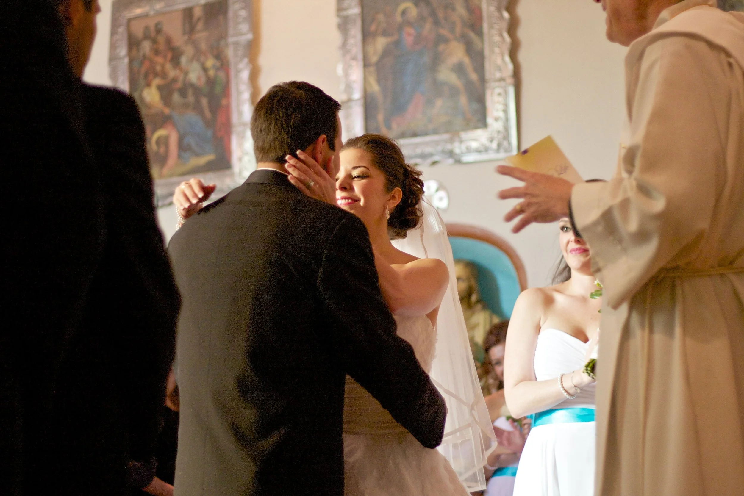 Bride and groom smiling and kissing during their wedding ceremony, surrounded by bridesmaids and officiant inside a decorated venue.
