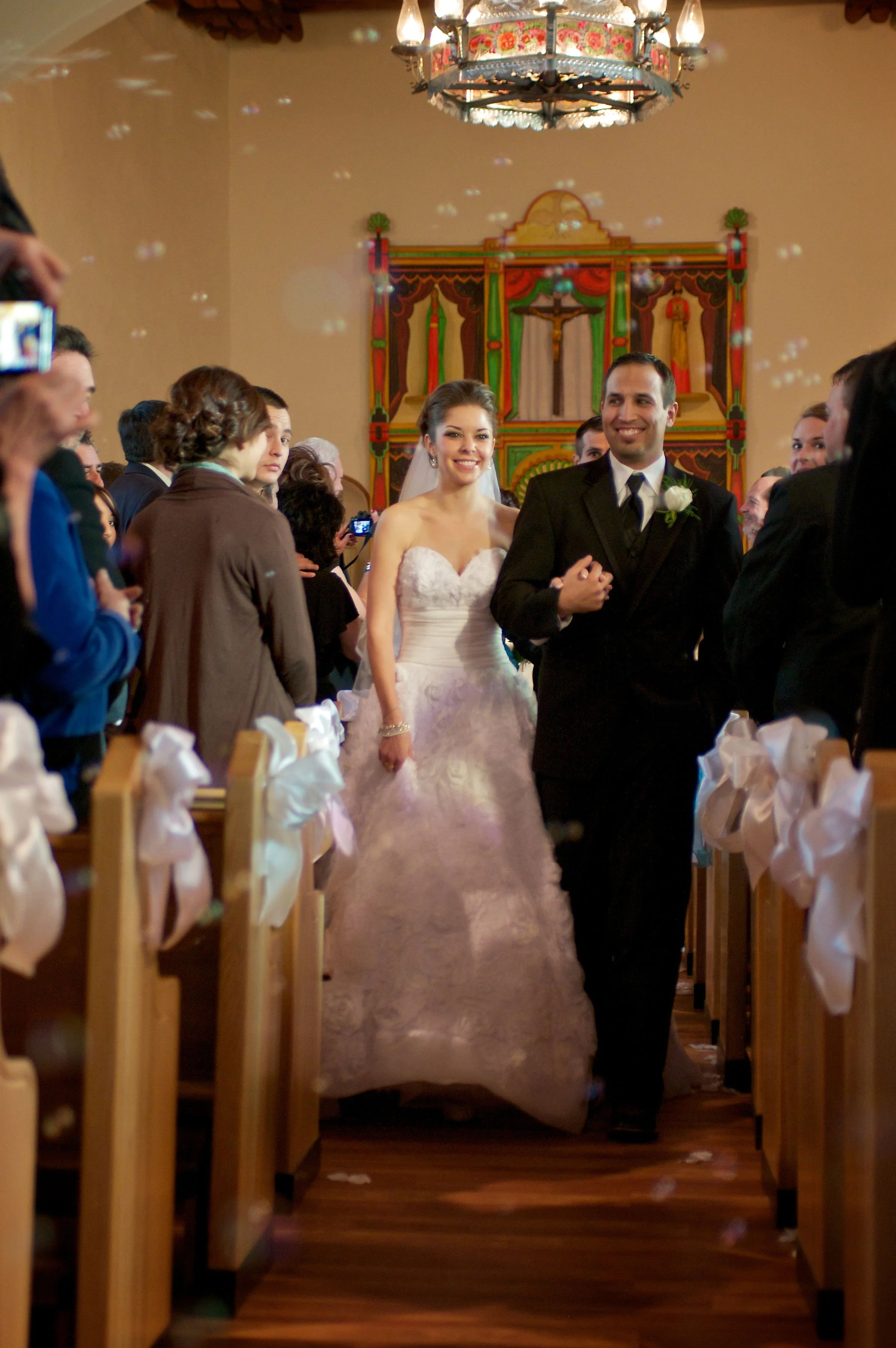 A bride in a white wedding dress and a groom in a black suit walking down the aisle of a church, smiling. Guests on both sides of the aisle watch and take photos. Church decor includes a colorful backdrop, candles, and bow decorations on the pews.