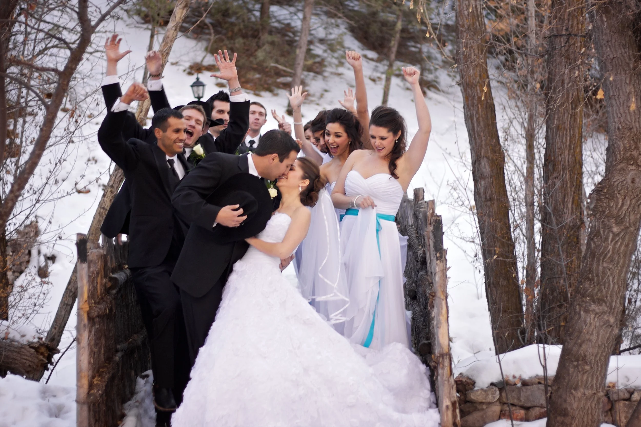Wedding party celebrating outdoors in a snowy forest, with the bride and groom kissing and the bridesmaids and groomsmen cheering around them.