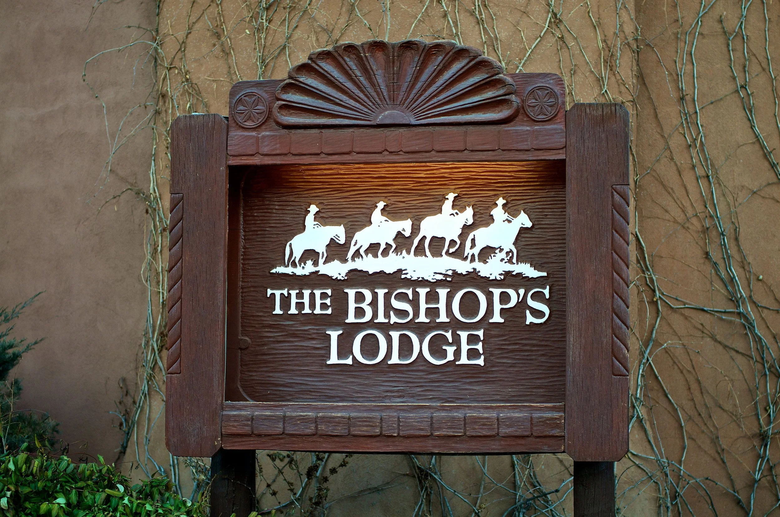 Wooden sign for the Bishop's Lodge featuring a graphic of five horses with riders and the text 'The Bishop's Lodge' below.