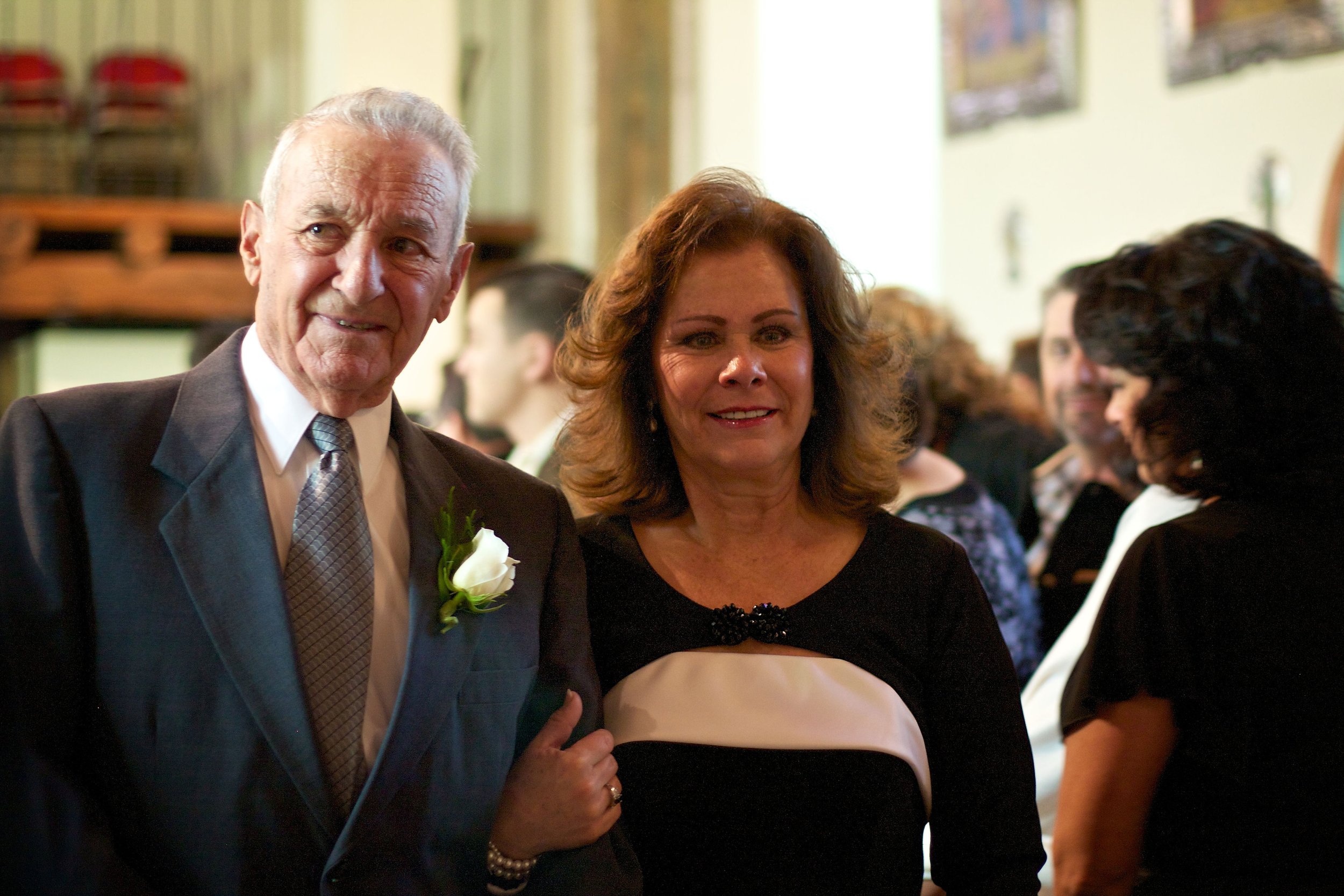 An elderly man in a suit wearing a white boutonniere stands next to a woman in a black and beige dress at a formal event with other people in the background.