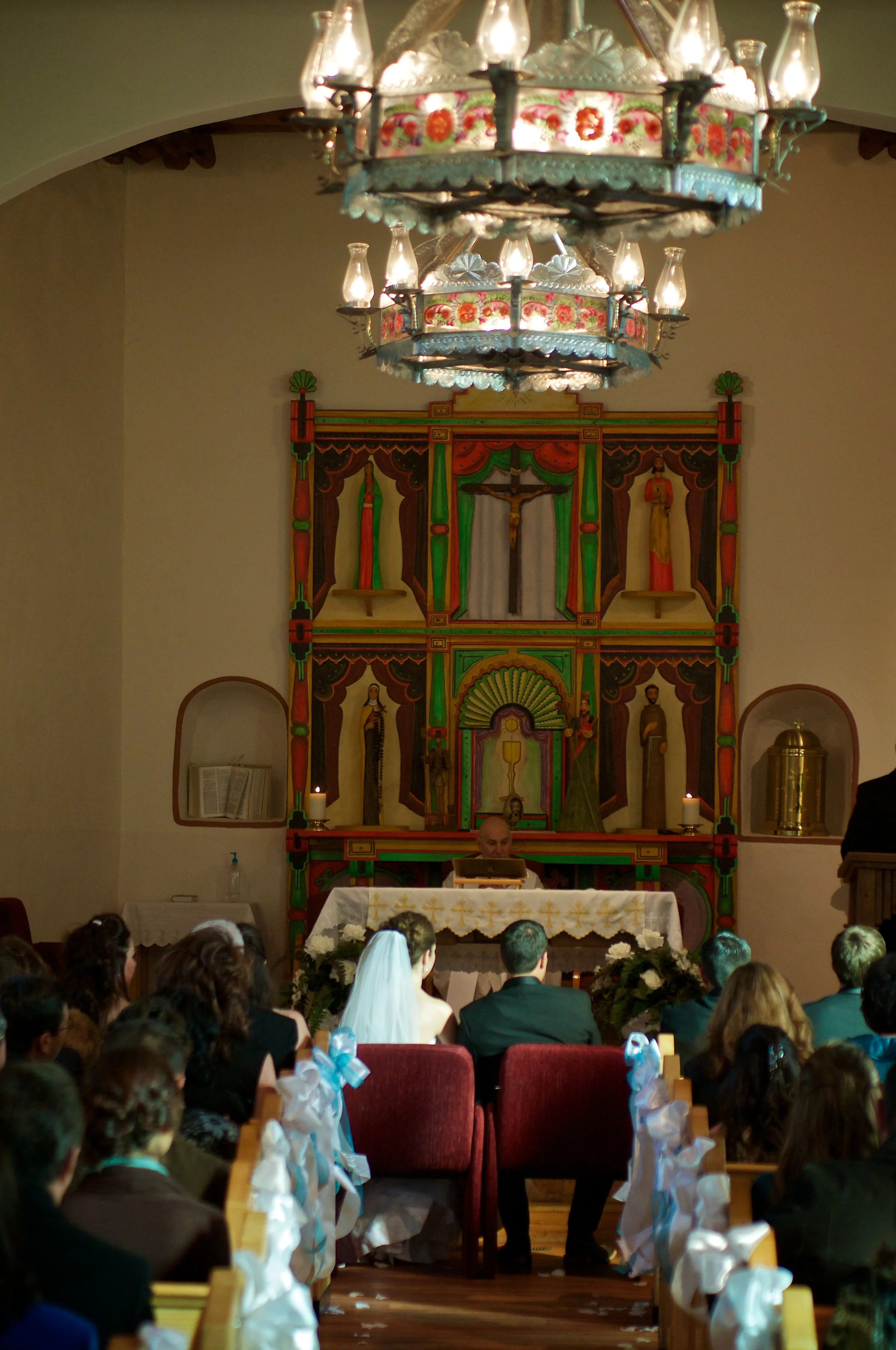 A wedding ceremony in a church with a priest behind an altar, a bride and groom seated, and guests facing forward, with traditional religious artwork and a chandelier overhead.