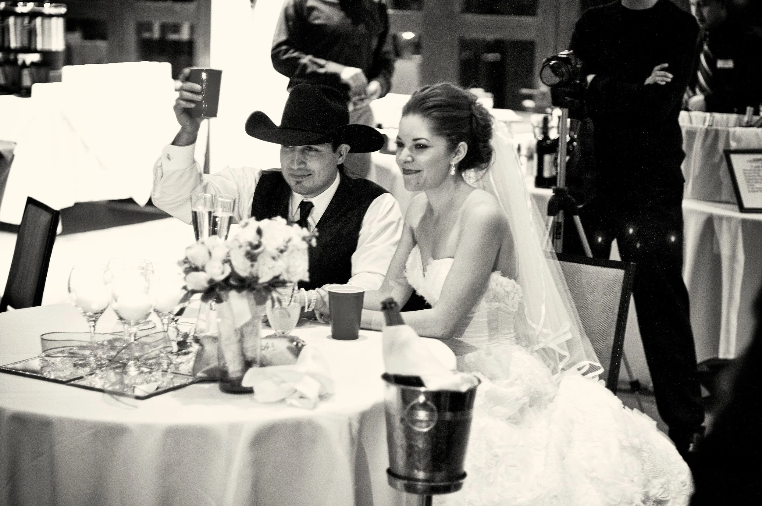 A bride and groom sit at a table during their wedding reception, with guests and photographers around them.