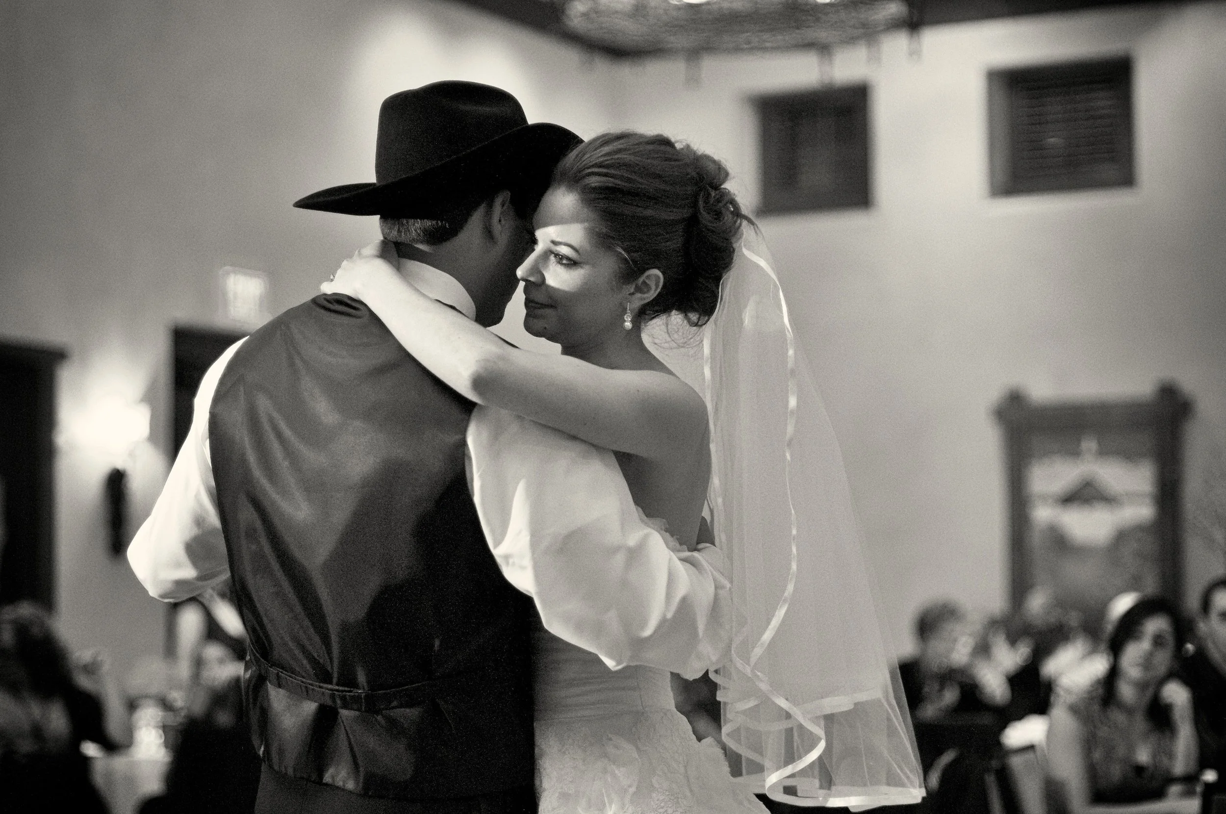 A black and white photo of a wedding dance with a bride and a groom, the bride is wearing a wedding dress and veil, and the groom is wearing a cowboy hat and vest, in a room with seated guests in the background.