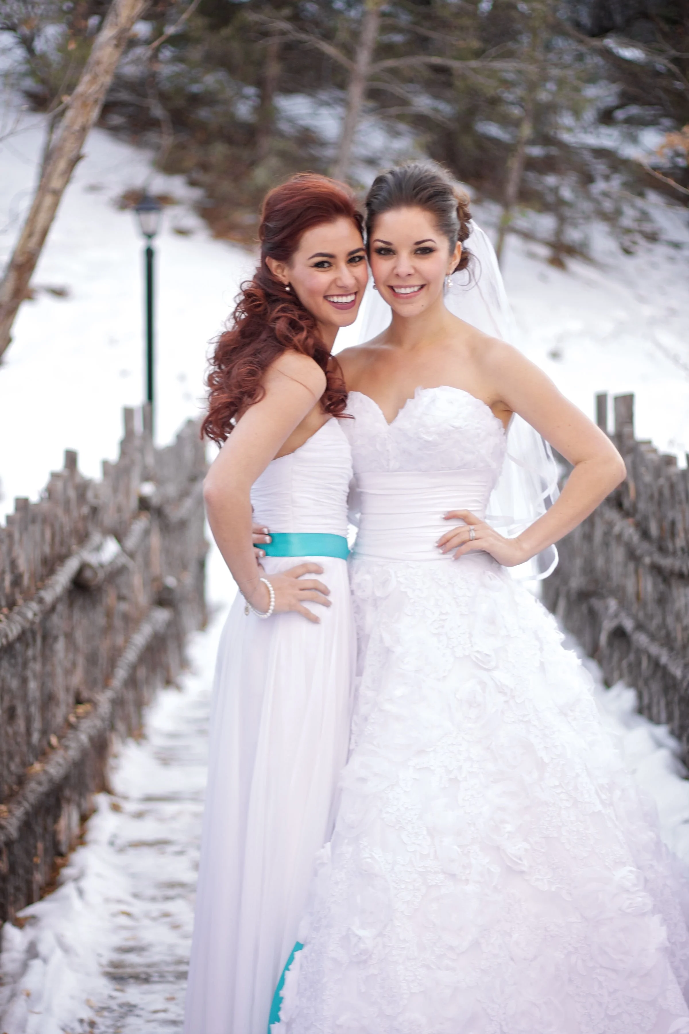 Two women in wedding dresses smiling on a snowy outdoor bridge, one with red hair and the other with brown hair, standing close together.
