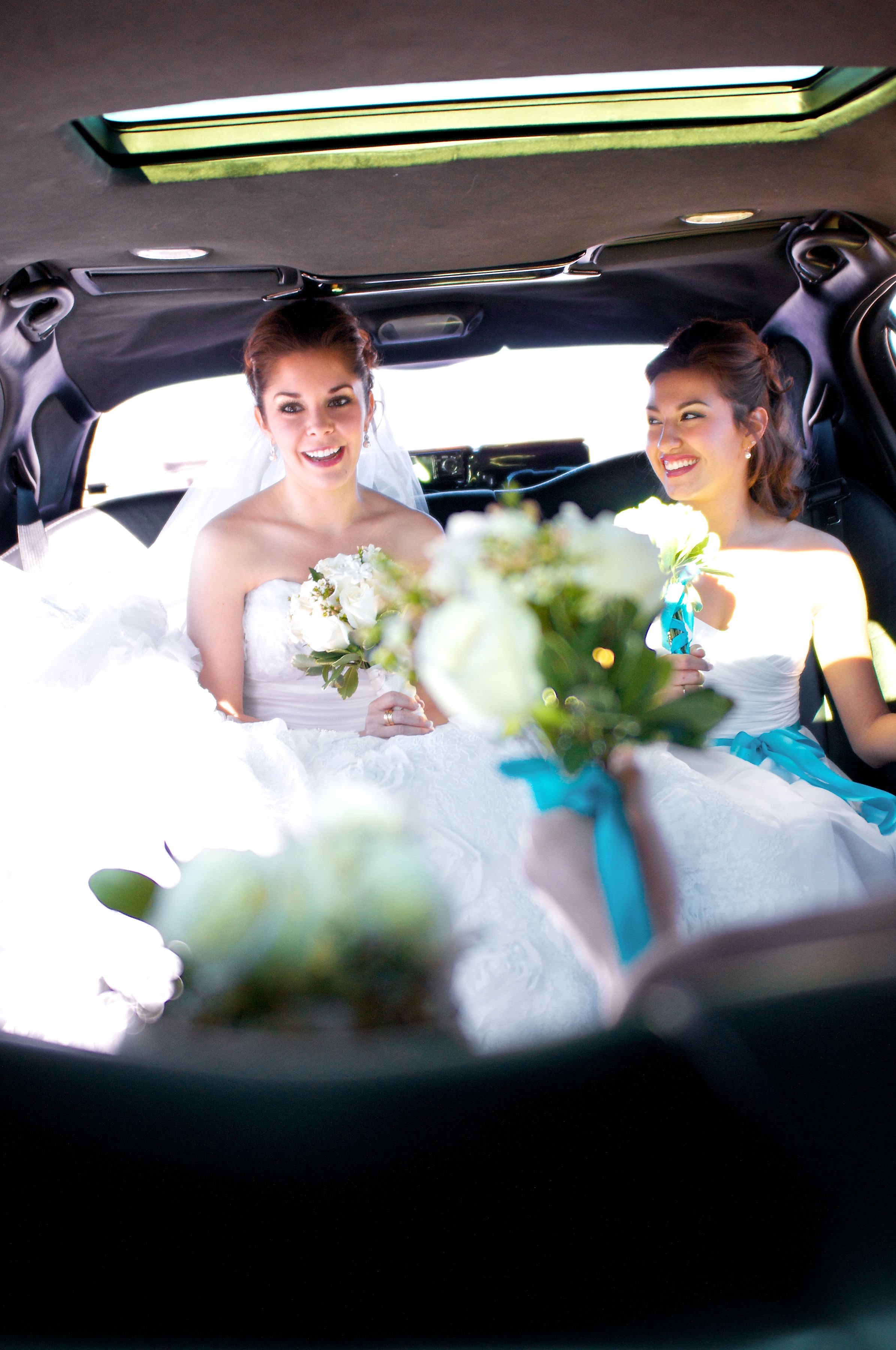 Bride in wedding dress sitting in limousine with bridesmaid, holding bouquets, with hands offering flowers in the foreground.