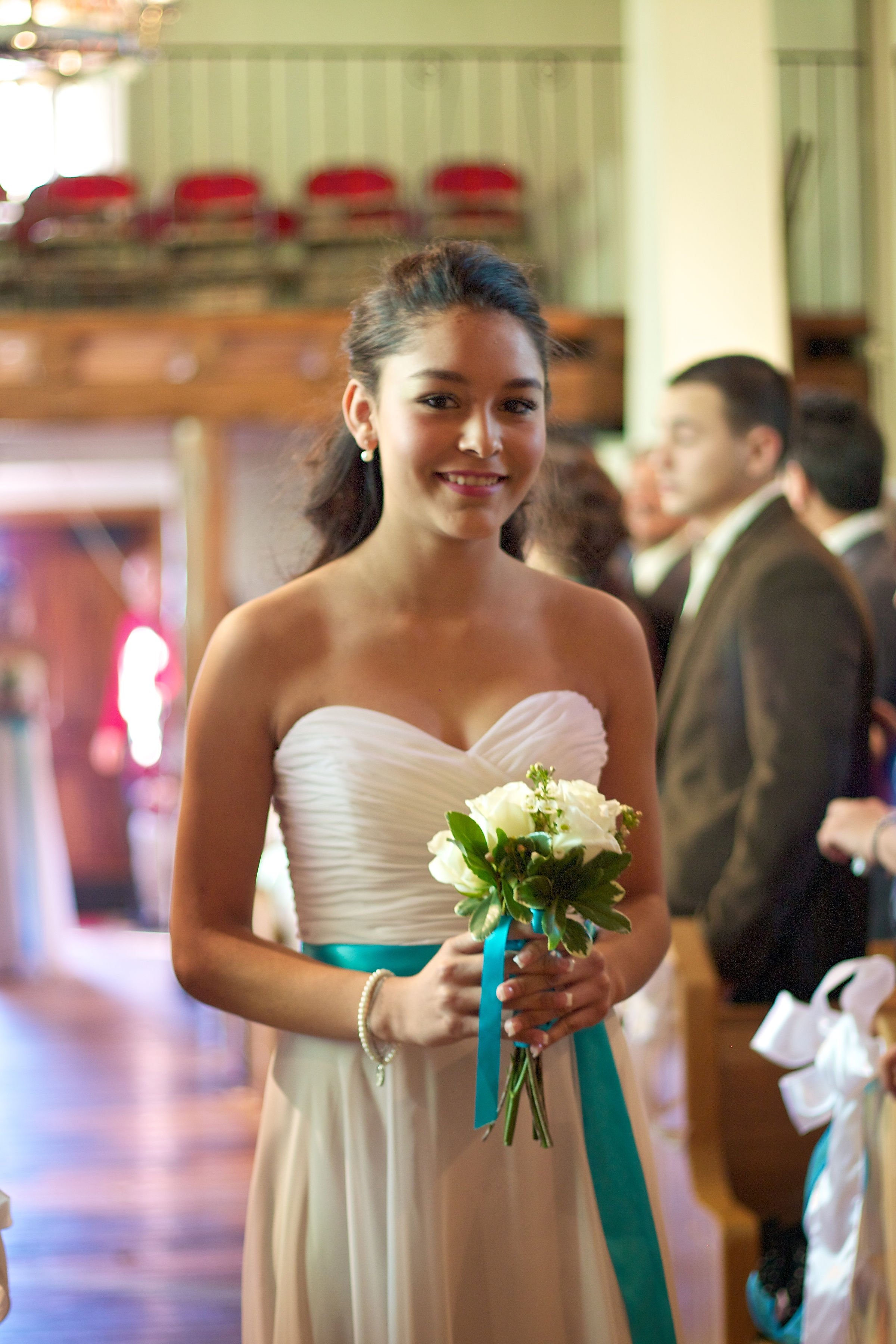 A young woman in a strapless white dress holding a small bouquet of white flowers, standing inside a decorated venue with other people in formal attire in the background.