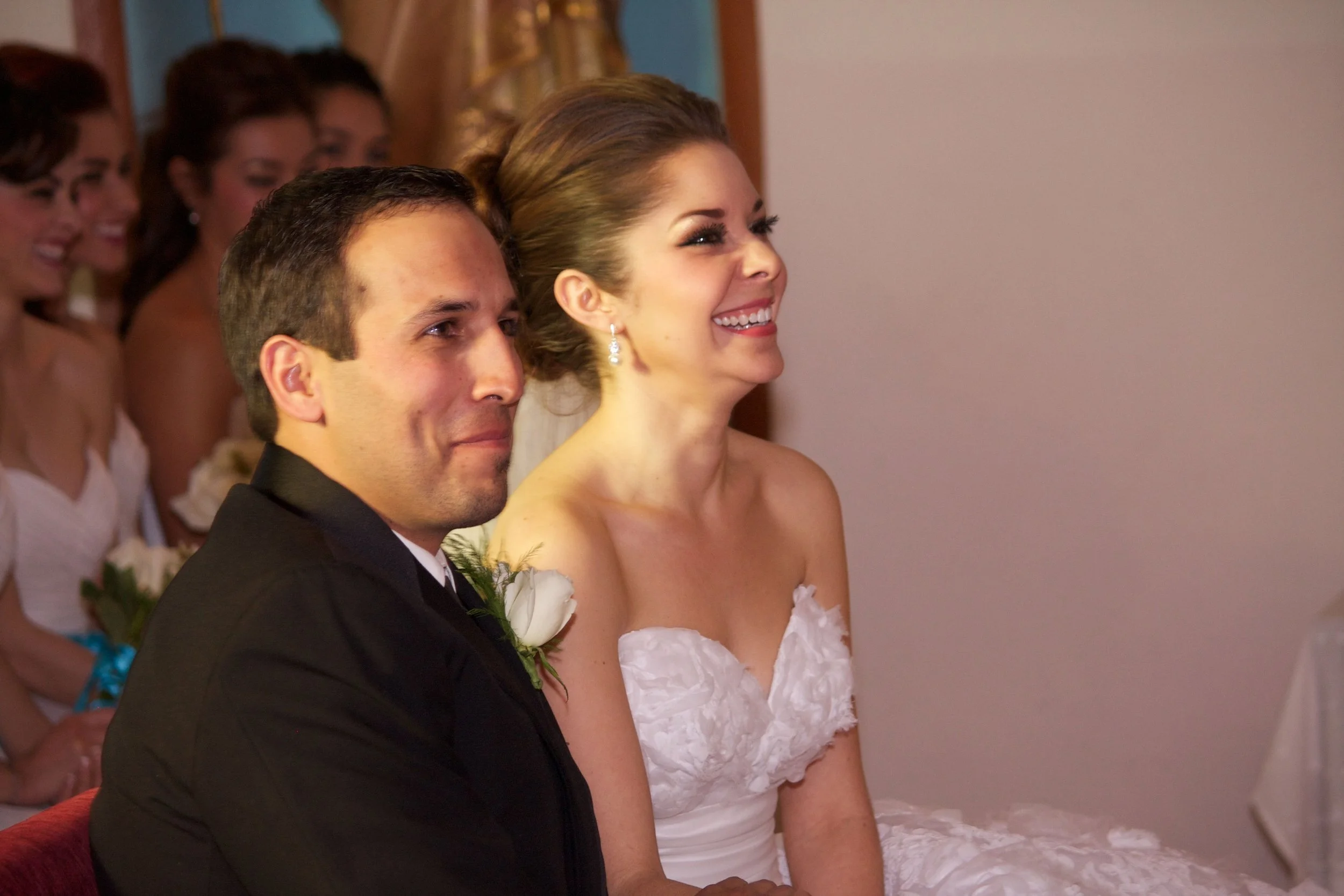 A bride and groom sitting together during their wedding ceremony, with wedding party members in the background.