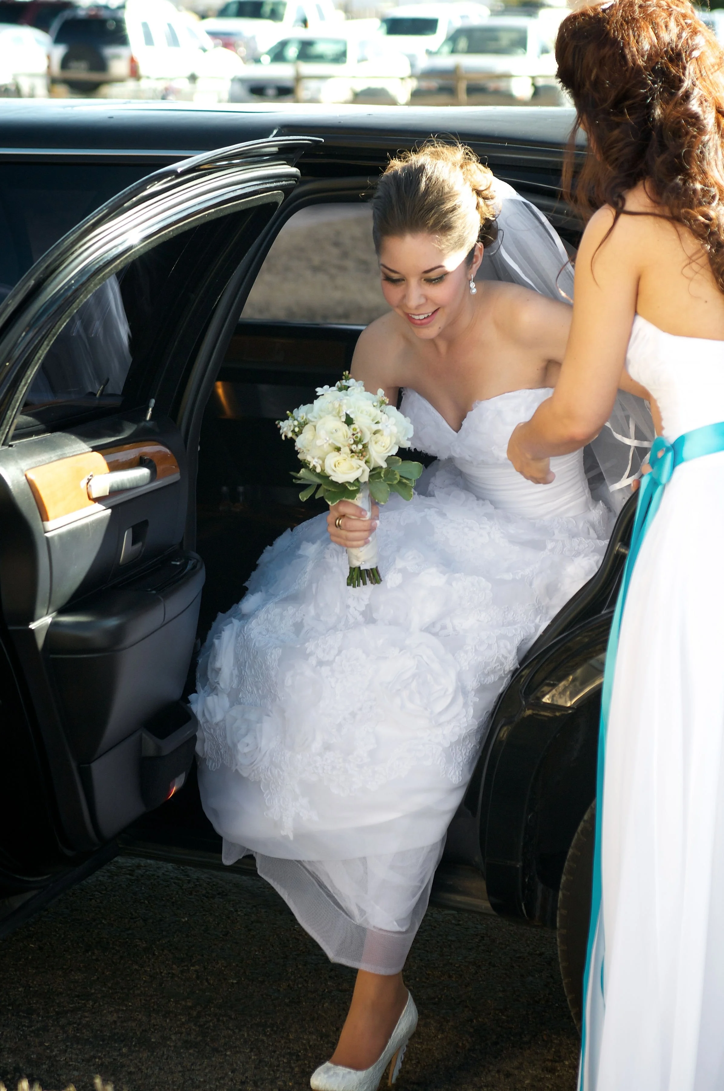 Bride in a wedding dress sitting in a car, holding a bouquet of white roses, while a woman helps her with her dress outside the car.