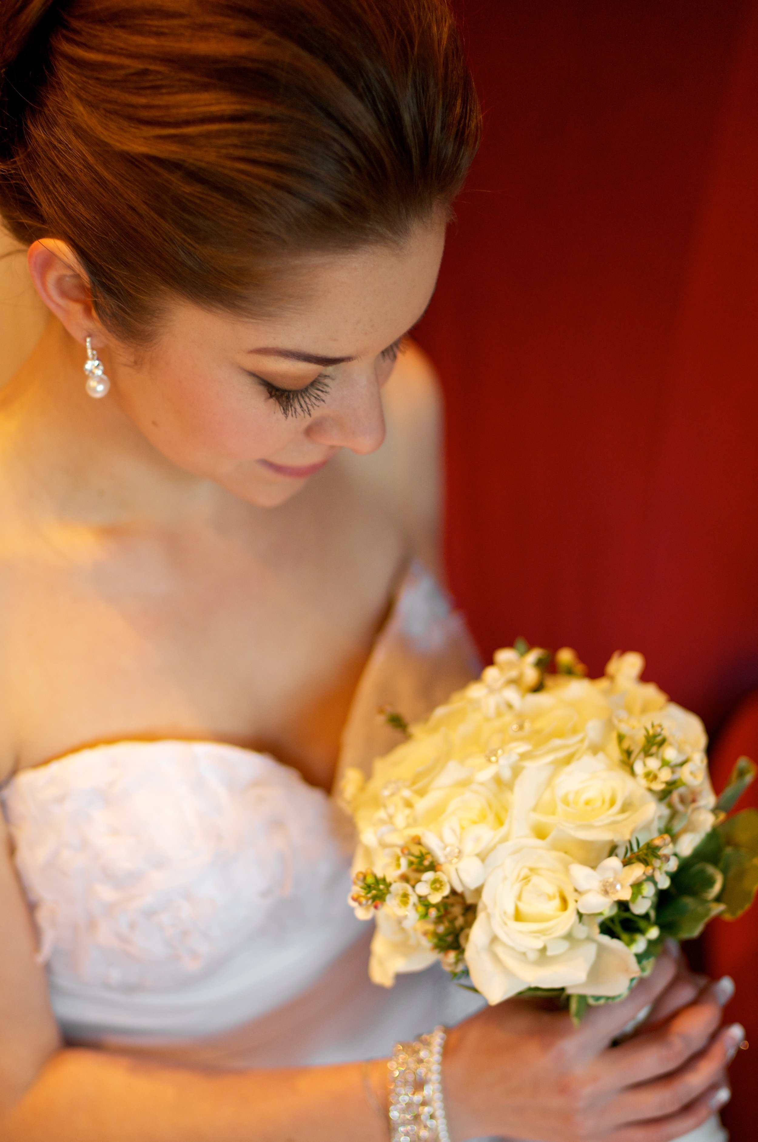 A bride with pearl earrings and a white strapless wedding dress holding a bouquet of white roses and small white flowers, looking down at her bouquet.