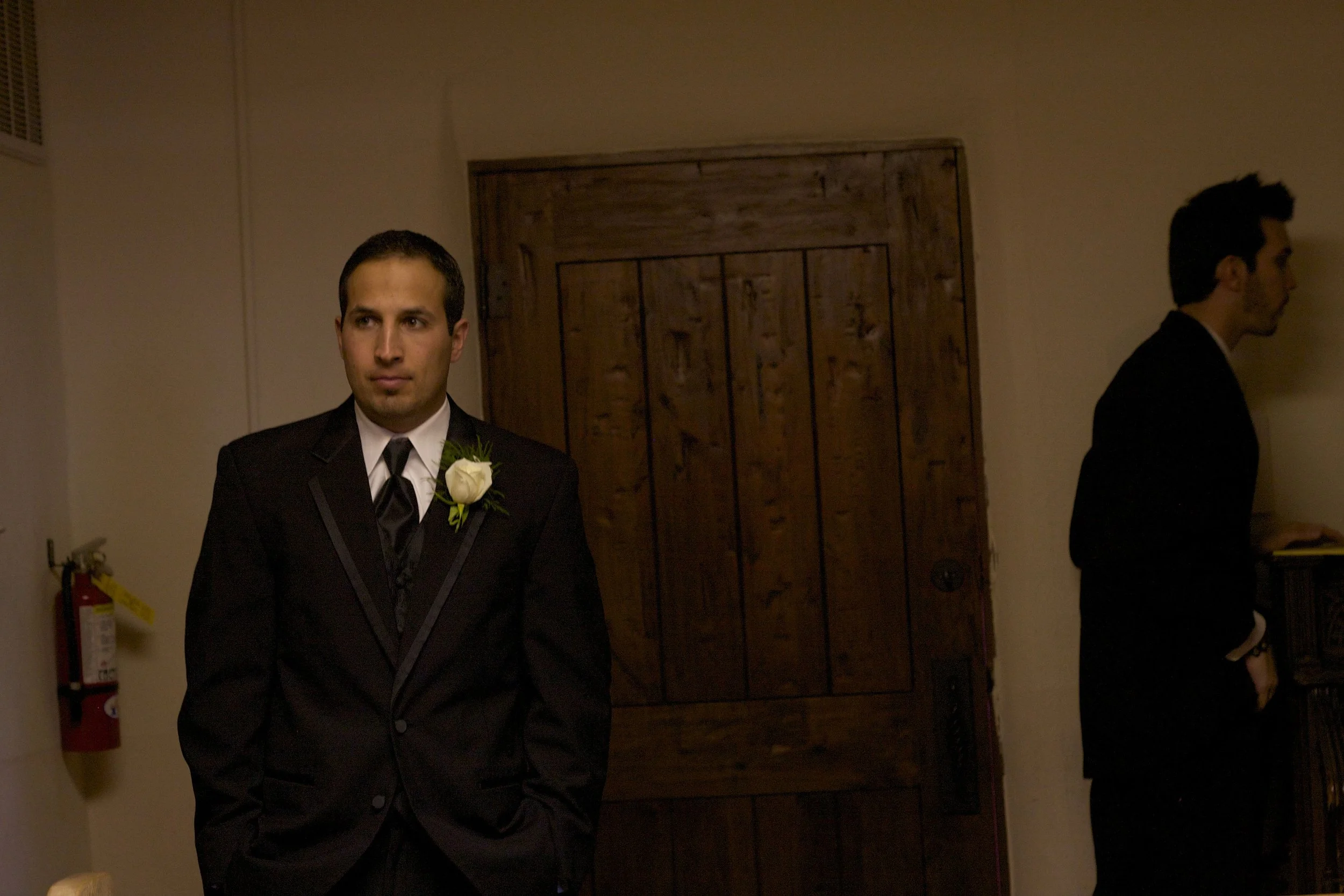 A man in a black suit with a white boutonniere stands with hands in his pockets, looking serious, in a dimly lit room. Another man in a black suit is turned sideways, looking away, near a wooden door.